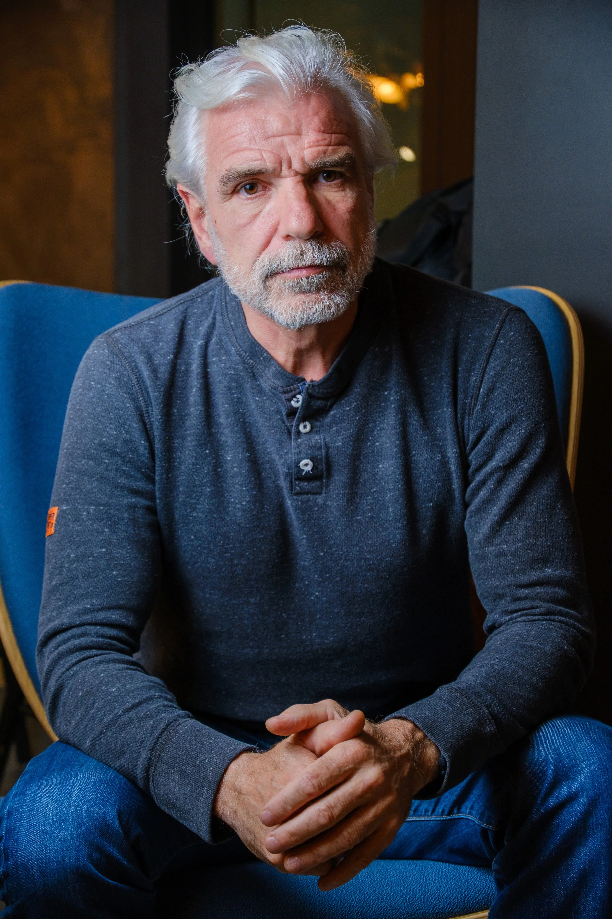 Homme âgé aux cheveux gris, barbe blanche, assis dans un fauteuil bleu, portant un pull gris foncé, regardant sérieusement vers l'objectif.