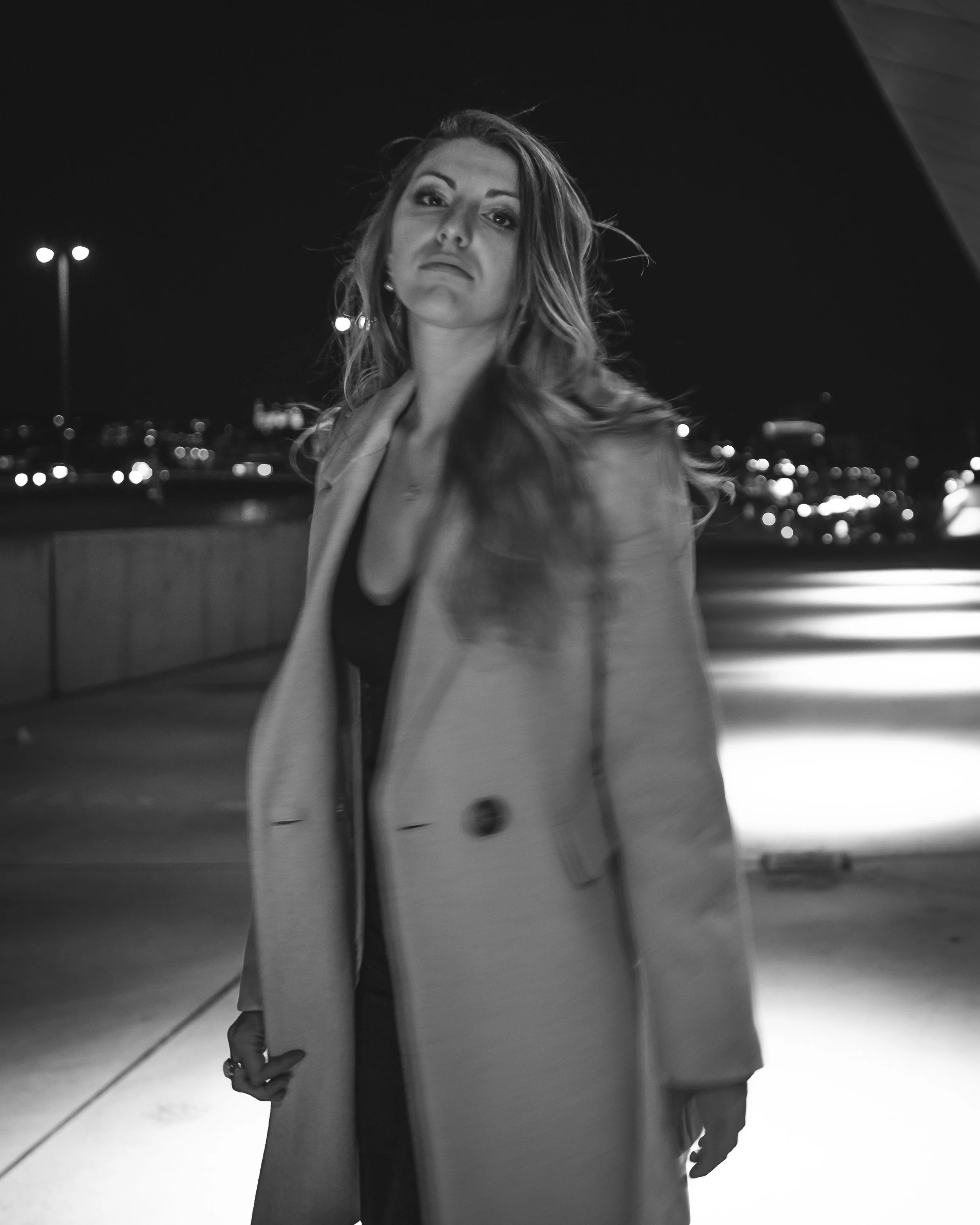 une femme debout la nuit, portant un trench beige, avec une ville en arrière-plan