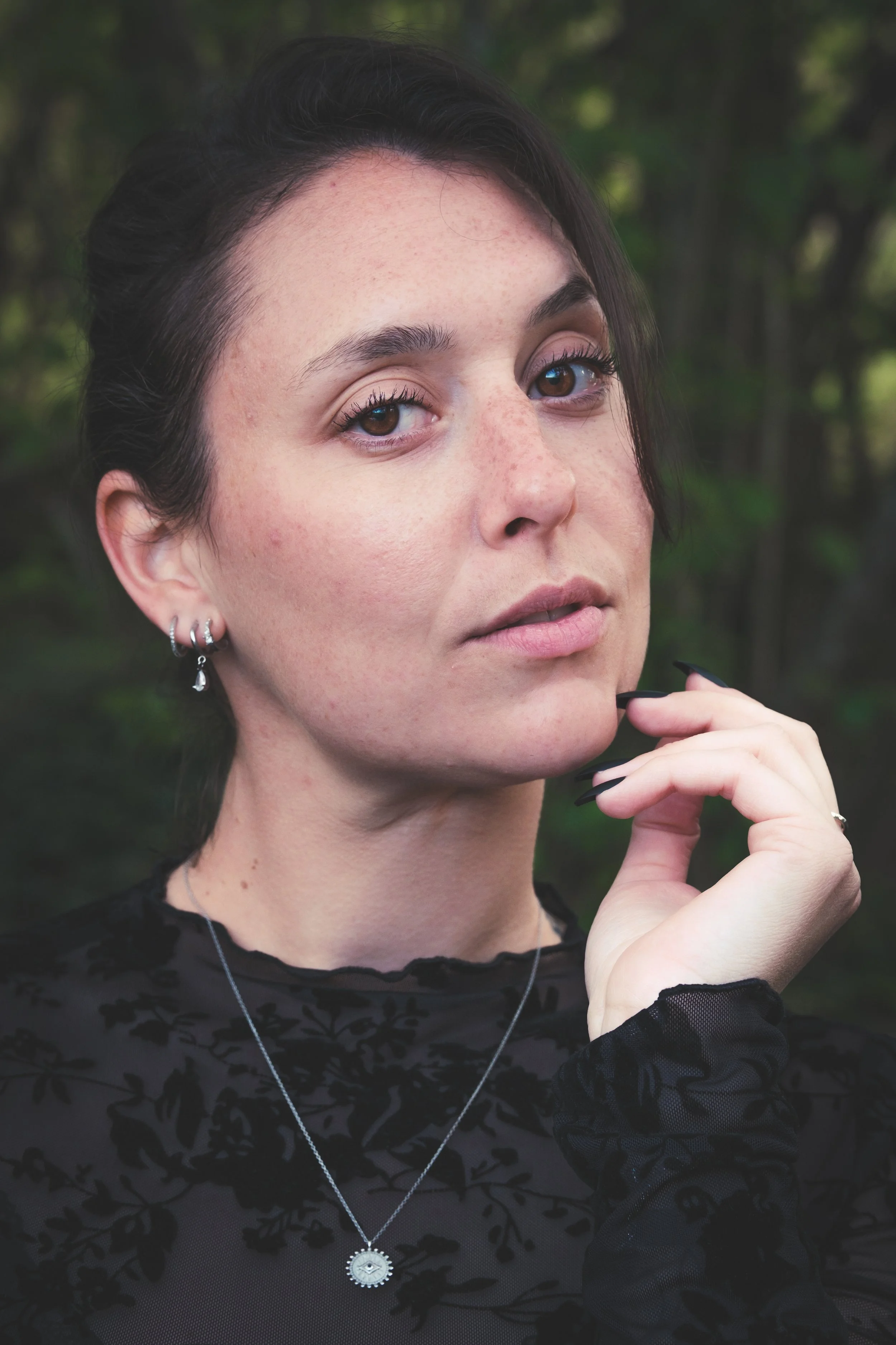 Jeune femme avec des cheveux noirs, maquillage léger, portant des bijoux en argent, une robe en voile noir avec un motif floral, posant en extérieur avec un fond de forêt verte.