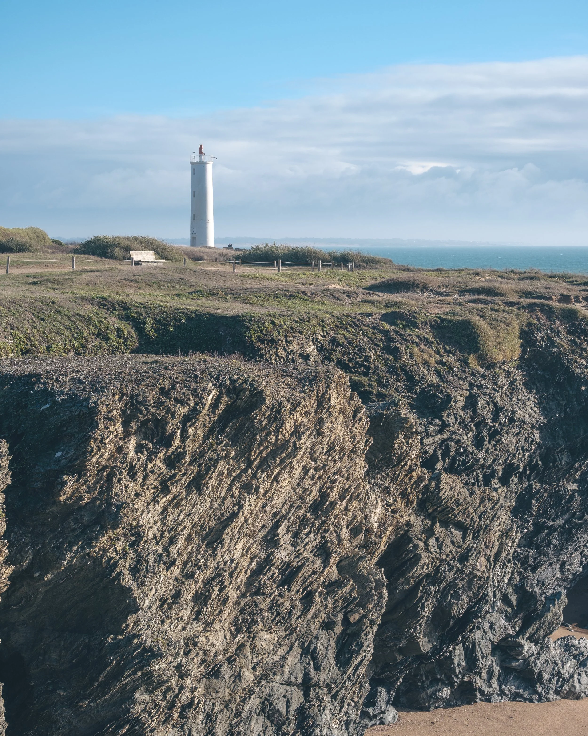 Phare blanc situé sur une falaise rocheuse au bord de l'océan avec un ciel partiellement nuageux.