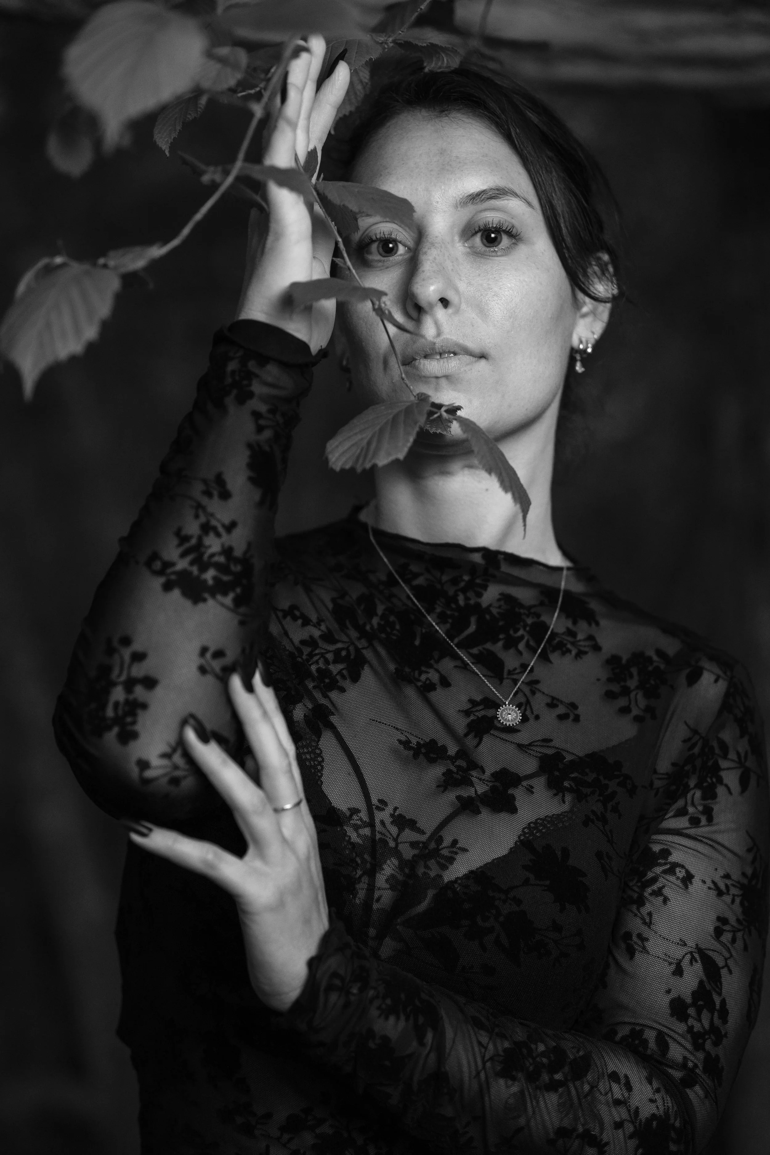 Une femme portant un vêtement en dentelle noire et une chaîne avec un pendentif. Elle tient une branche d'arbre devant son visage dans une photographie en noir et blanc.