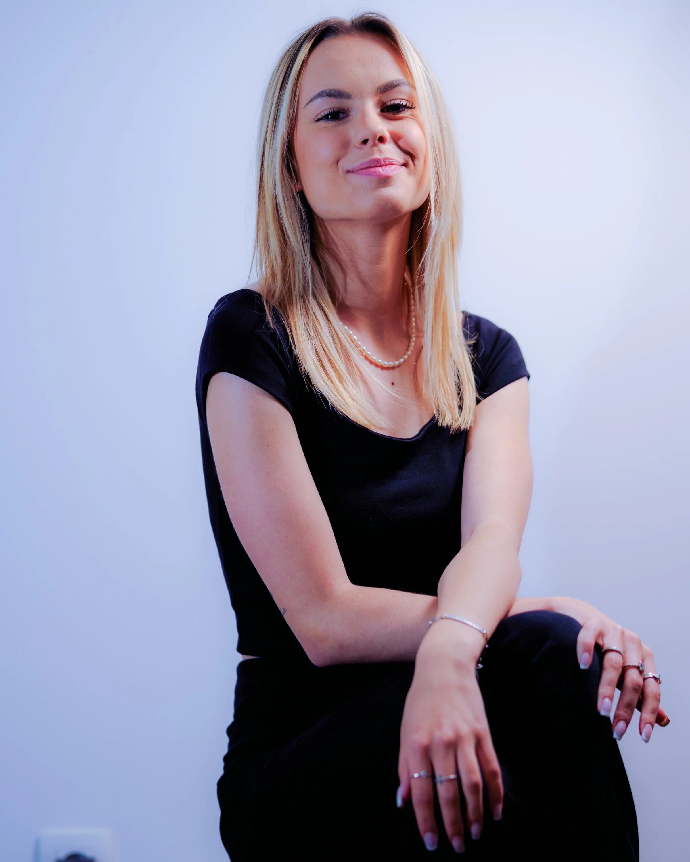 Une femme blonde souriante portant un collier de perles, vêtue en noir, assise contre un fond blanc