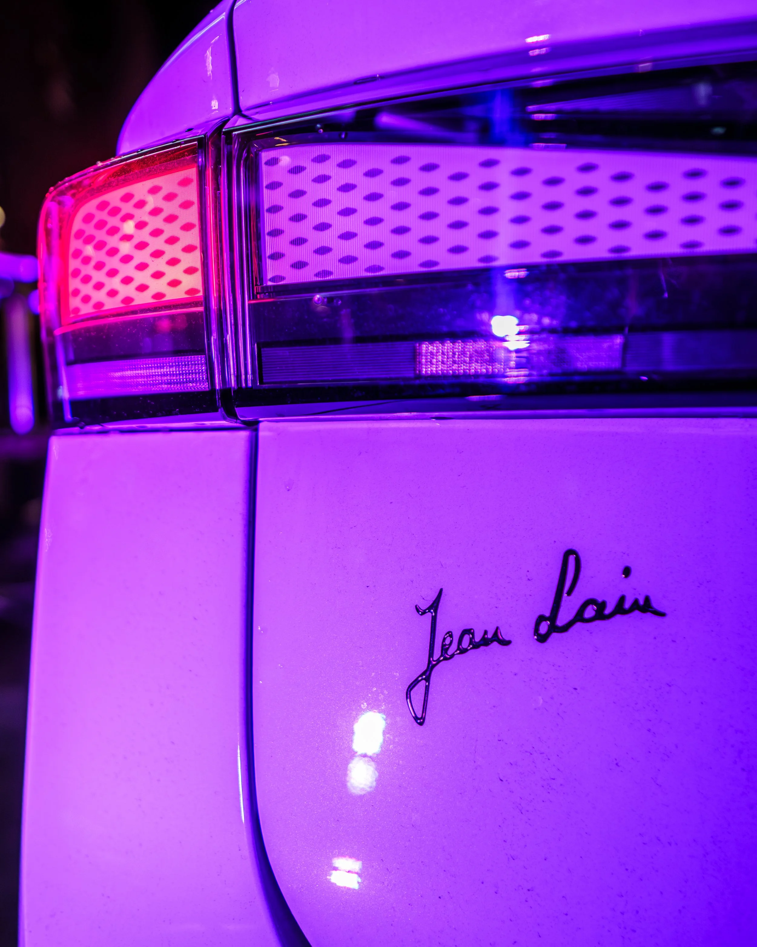 Détail d'une partie arrière d'une voiture blanche avec un éclairage violet, mettant en valeur la lampe et la signature 'Jean Lain' sur la carrosserie.