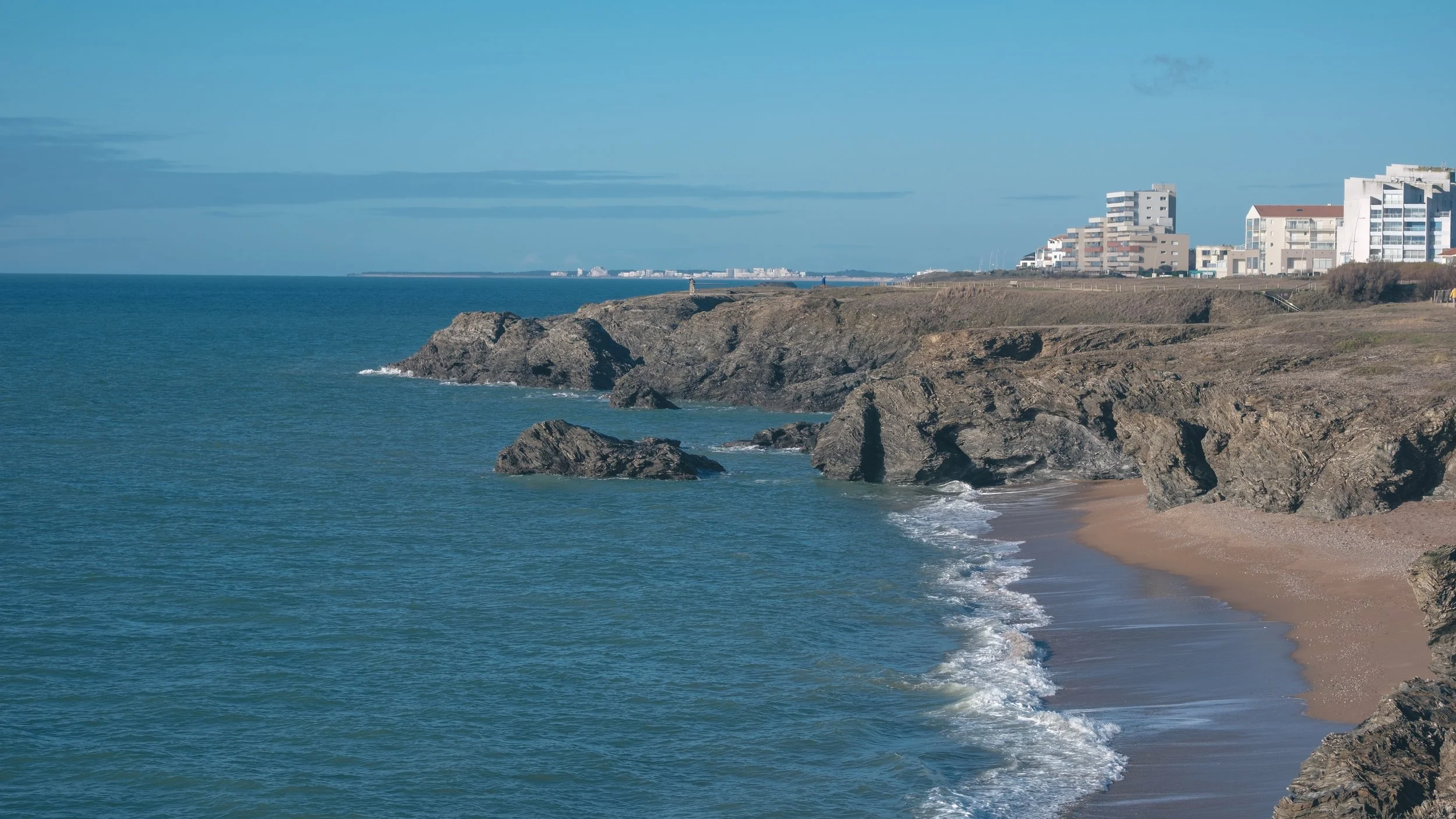 Côte rocheuse avec une plage de sable, bâtiments modernes en arrière-plan, océan clair sous un ciel bleu.