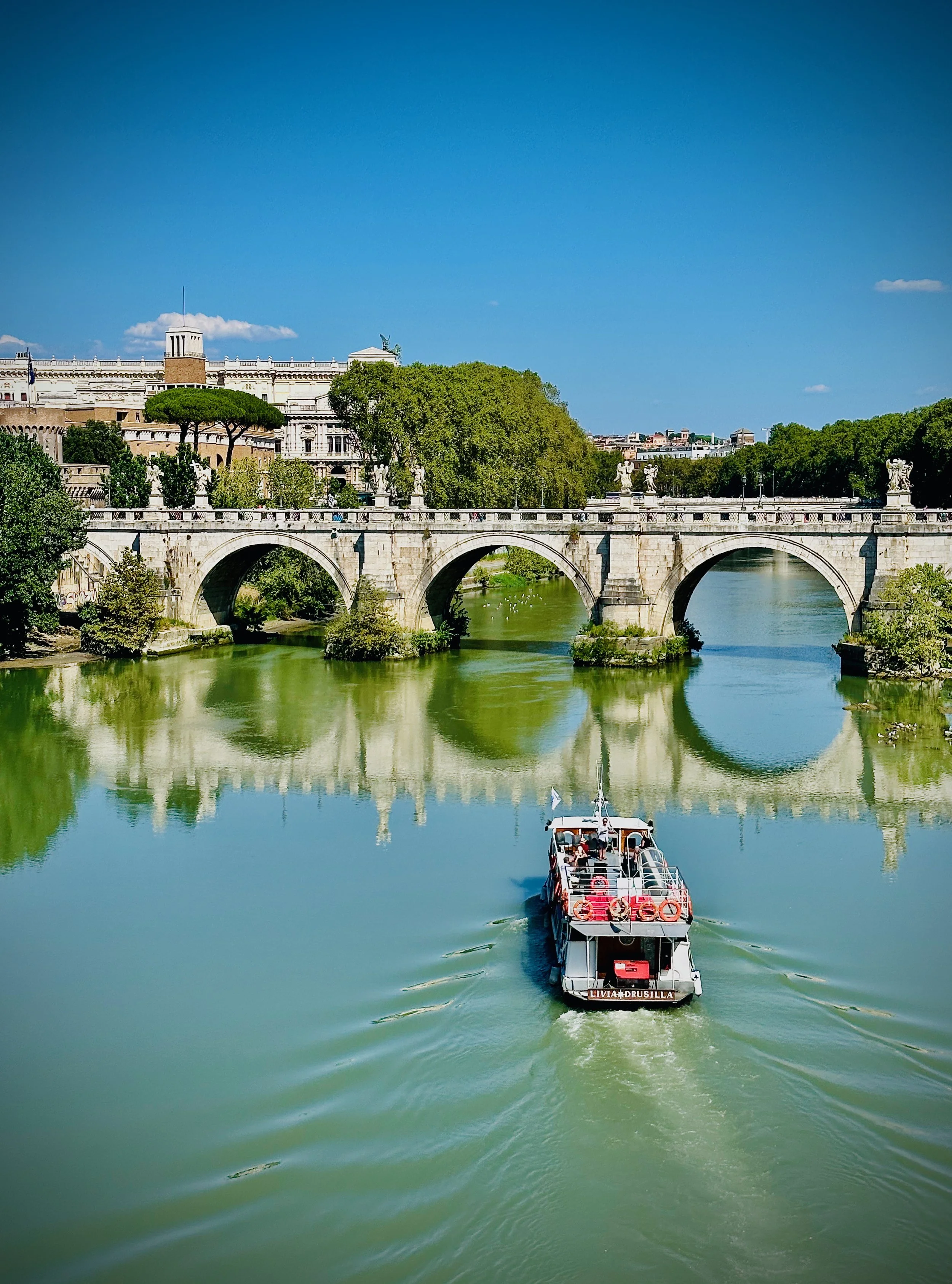 Un bateau navigue sur la rivière Tibre à Rome, avec un pont orné de statues en arrière-plan et un ciel bleu clair.