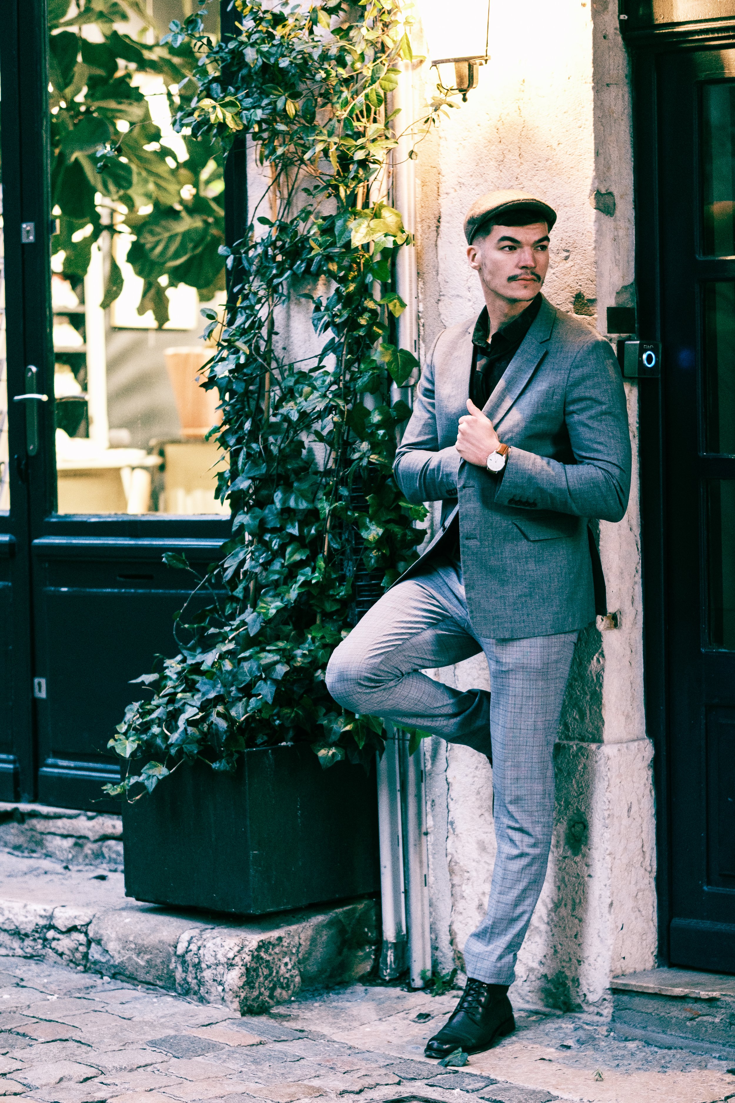 Un homme en costume gris, portant une montre, en train de poser contre un mur dans une rue pavée, avec une porte en bois à côté et une plante verte dans un pot noir.