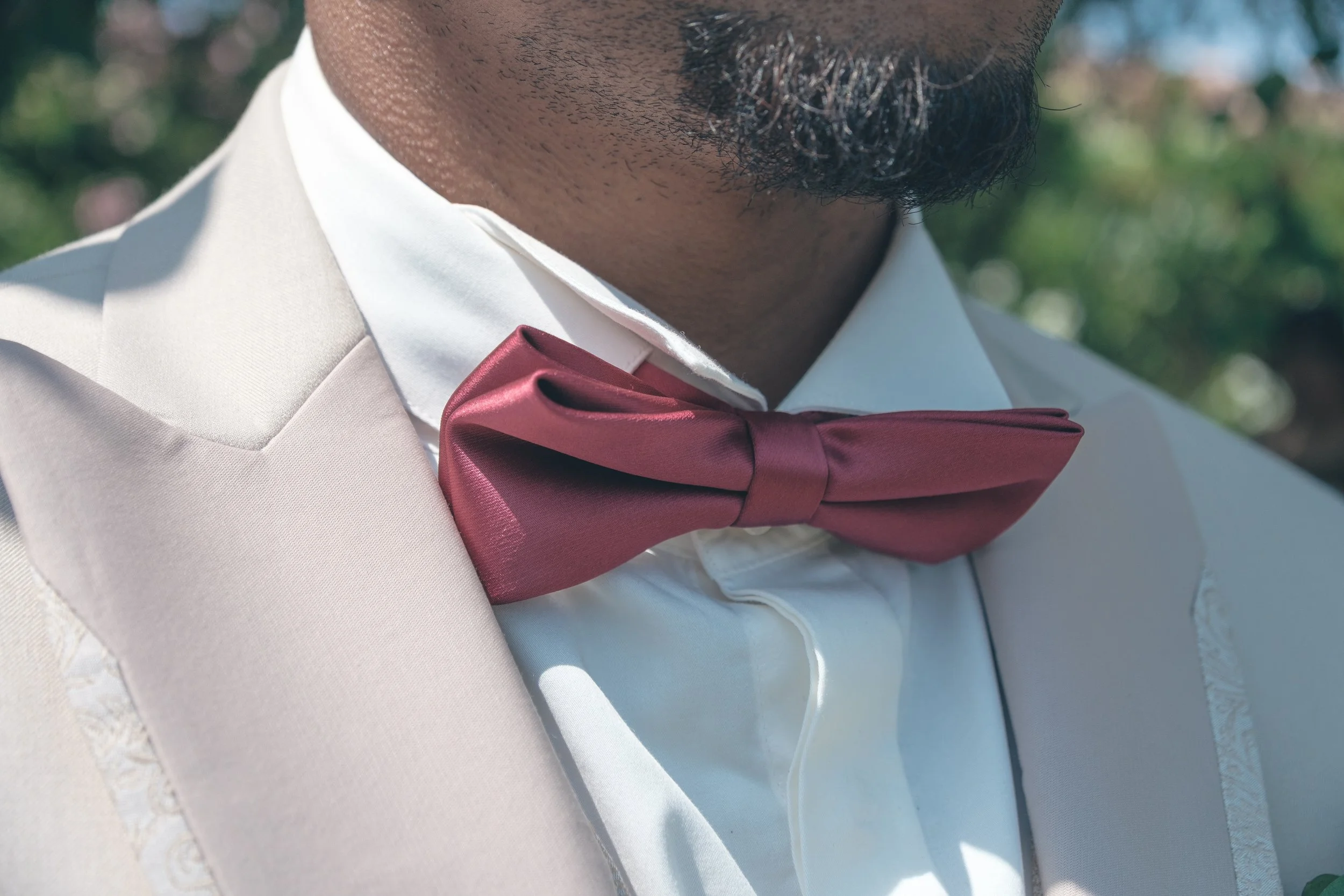Un homme portant un costume blanc avec une chemise blanche et un nœud papillon en satin rouge foncé, en plein air avec un fond flou de verdure.