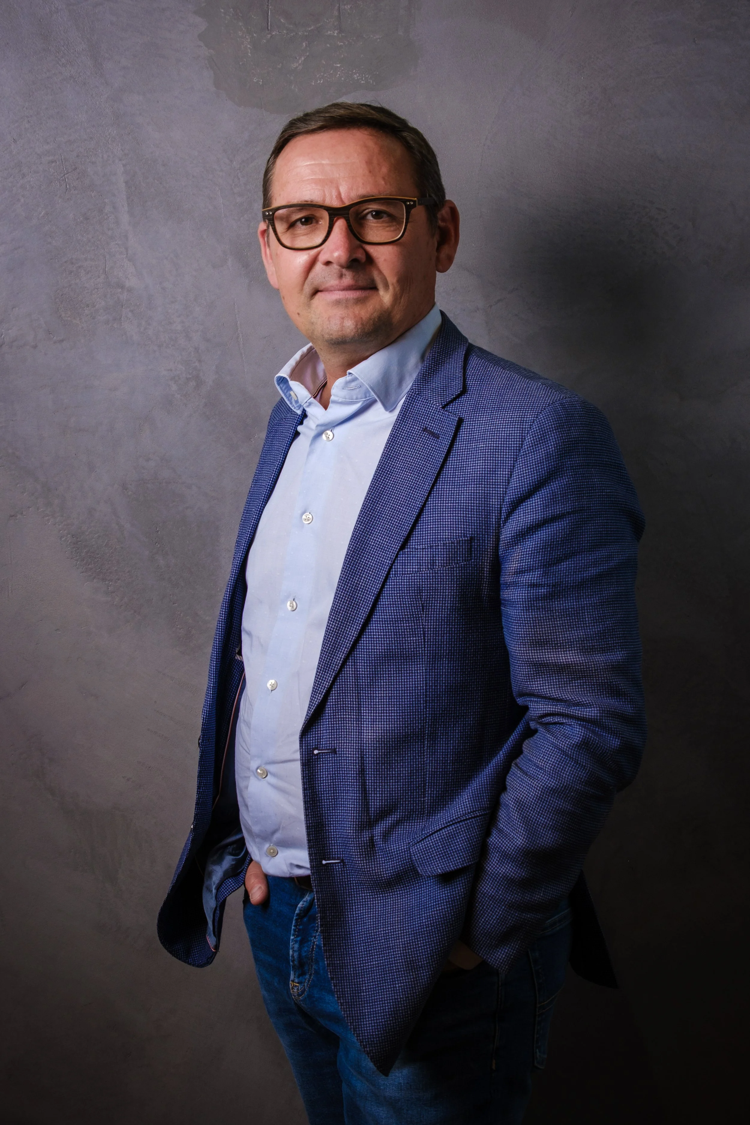 Homme adulte portant des lunettes, en blazer bleu à carreaux, chemise blanche, pantalon en jeans, posé contre un mur gris