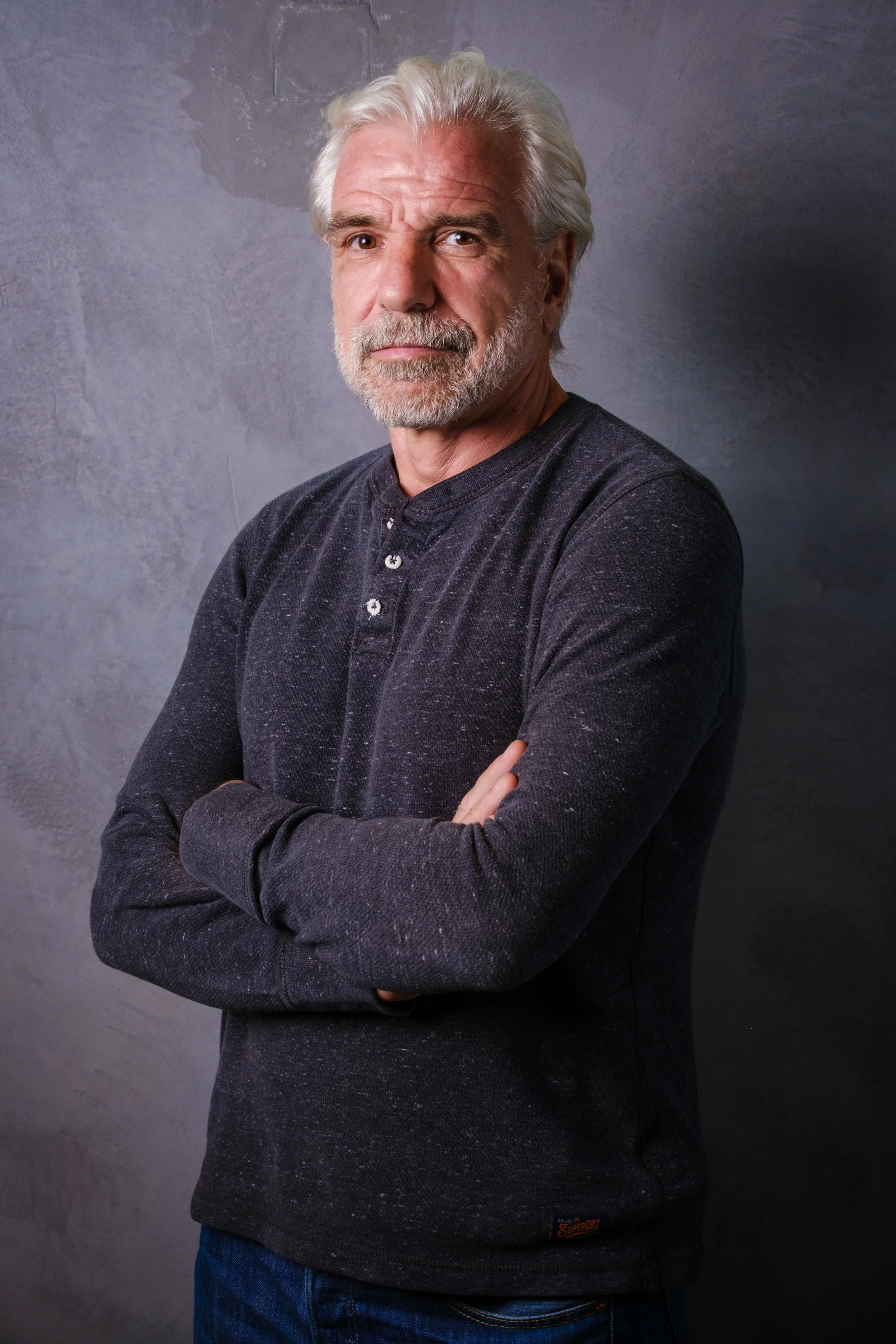 Un homme âgé aux cheveux gris et à la barbe, portant un pull noir à manches longues et croisés, pose avec les bras croisés devant un mur gris.