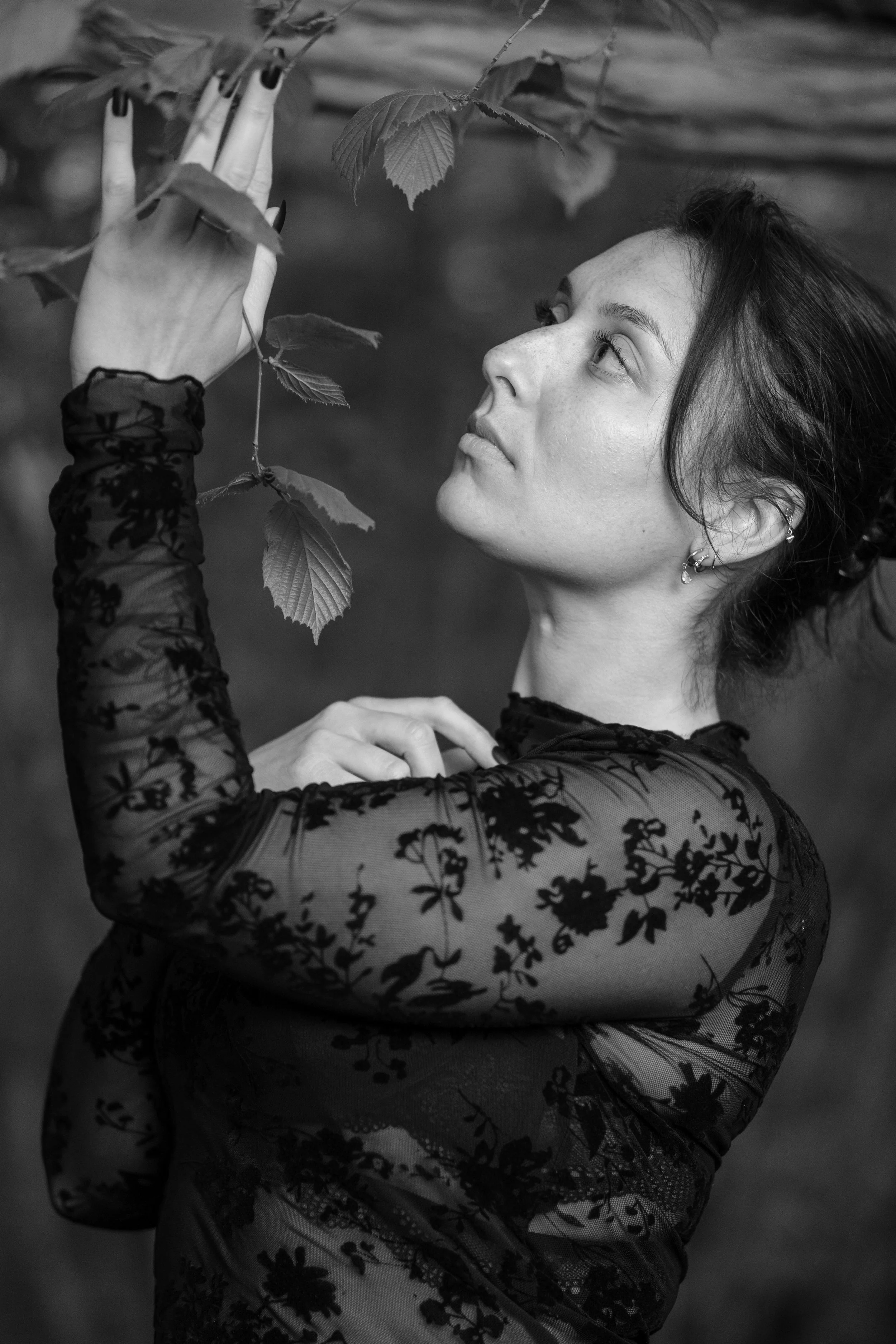 Une femme en robe en dentelle noire se tient près de branches d'arbre, regardant attentivement les feuilles.