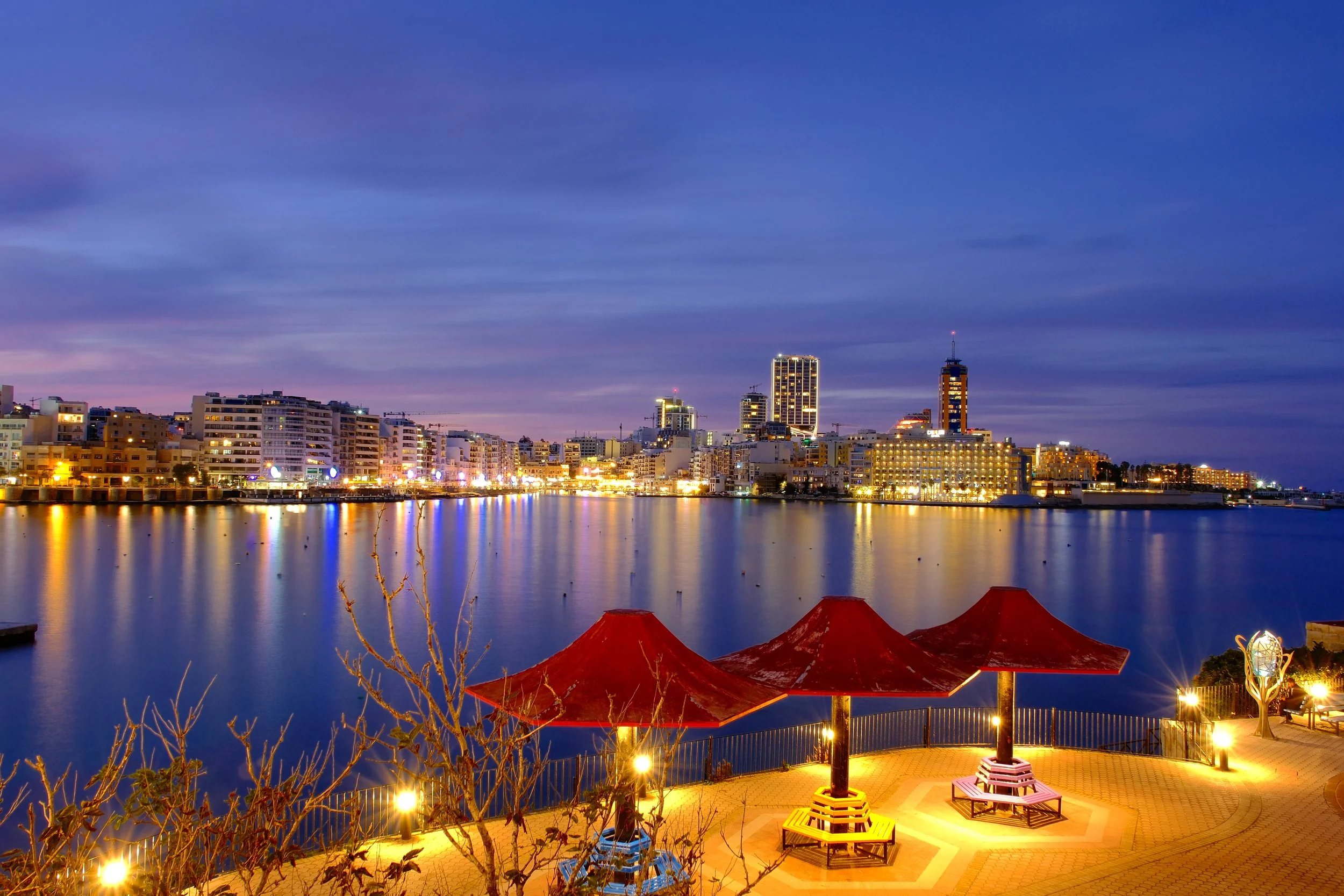 Vue nocturne d'une ville au bord de l'eau avec des bâtiments illuminés et des parasols rouges sur une terrasse.