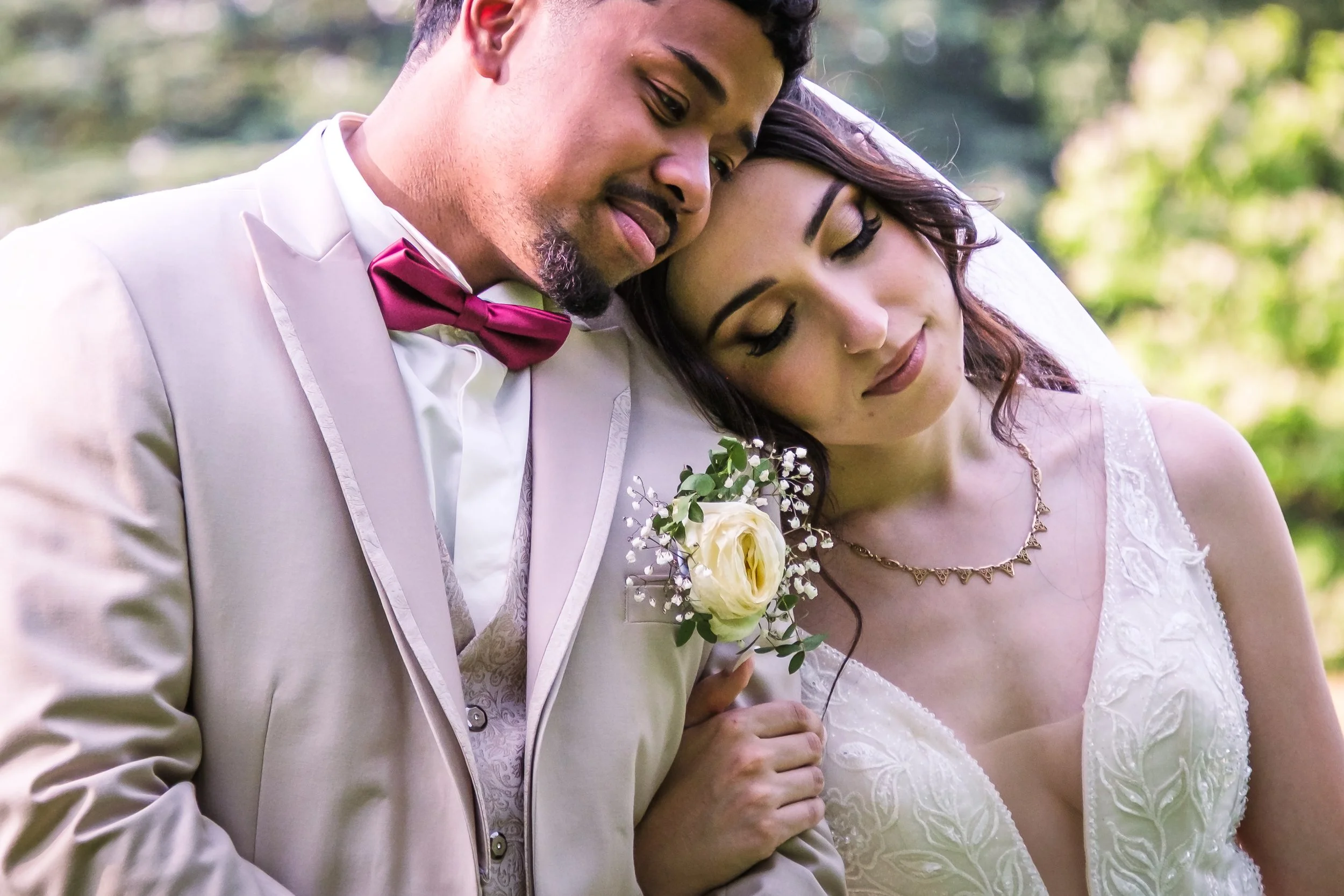 Un couple de mariés en robe de mariage et costume blanc, en train de se tenir proche, dans un jardin avec des arbres en arrière-plan.