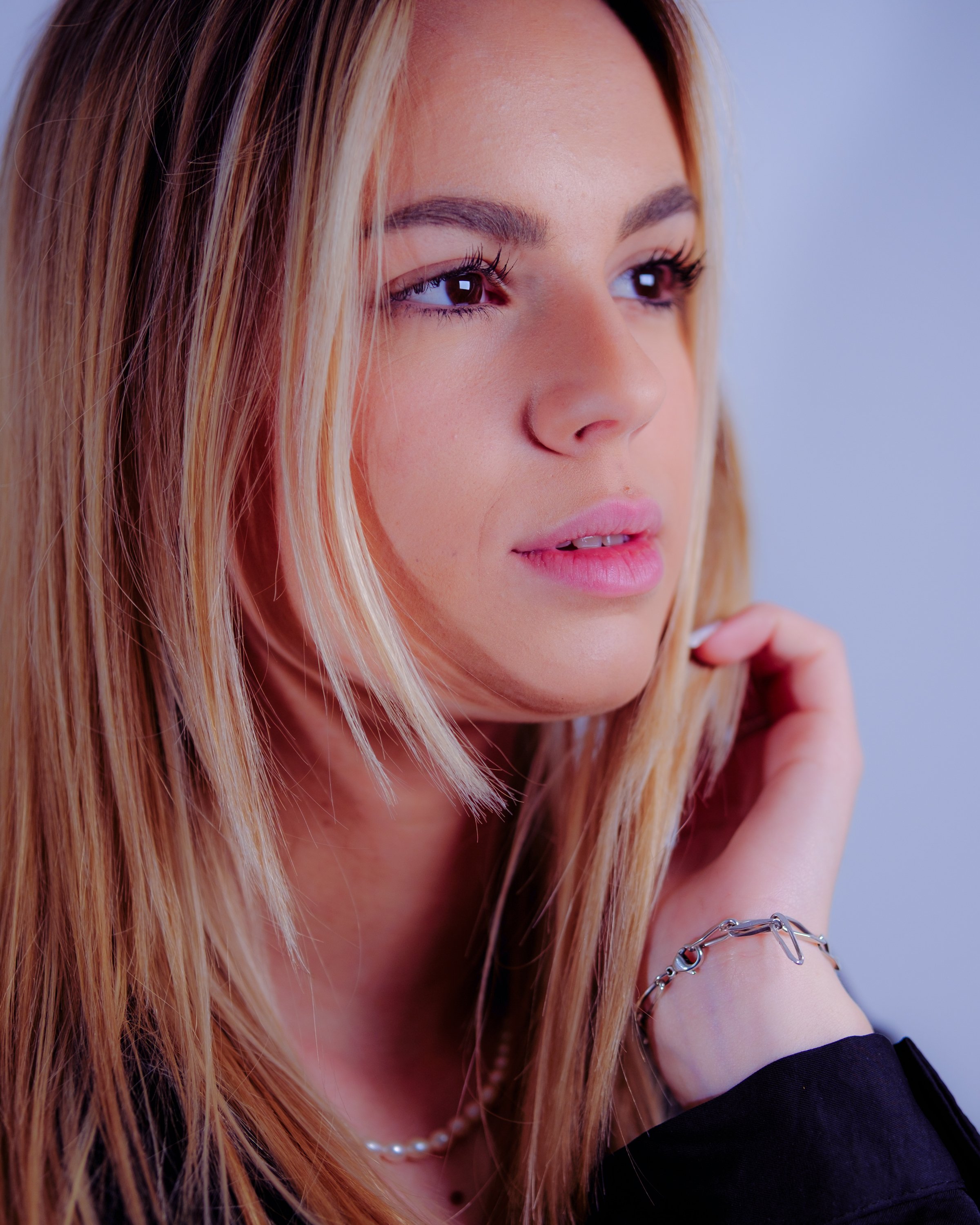 Jeune femme aux cheveux longs et blonds, portant une chaîne de perles et un bracelet en argent, paysage serein, expression contemplative.