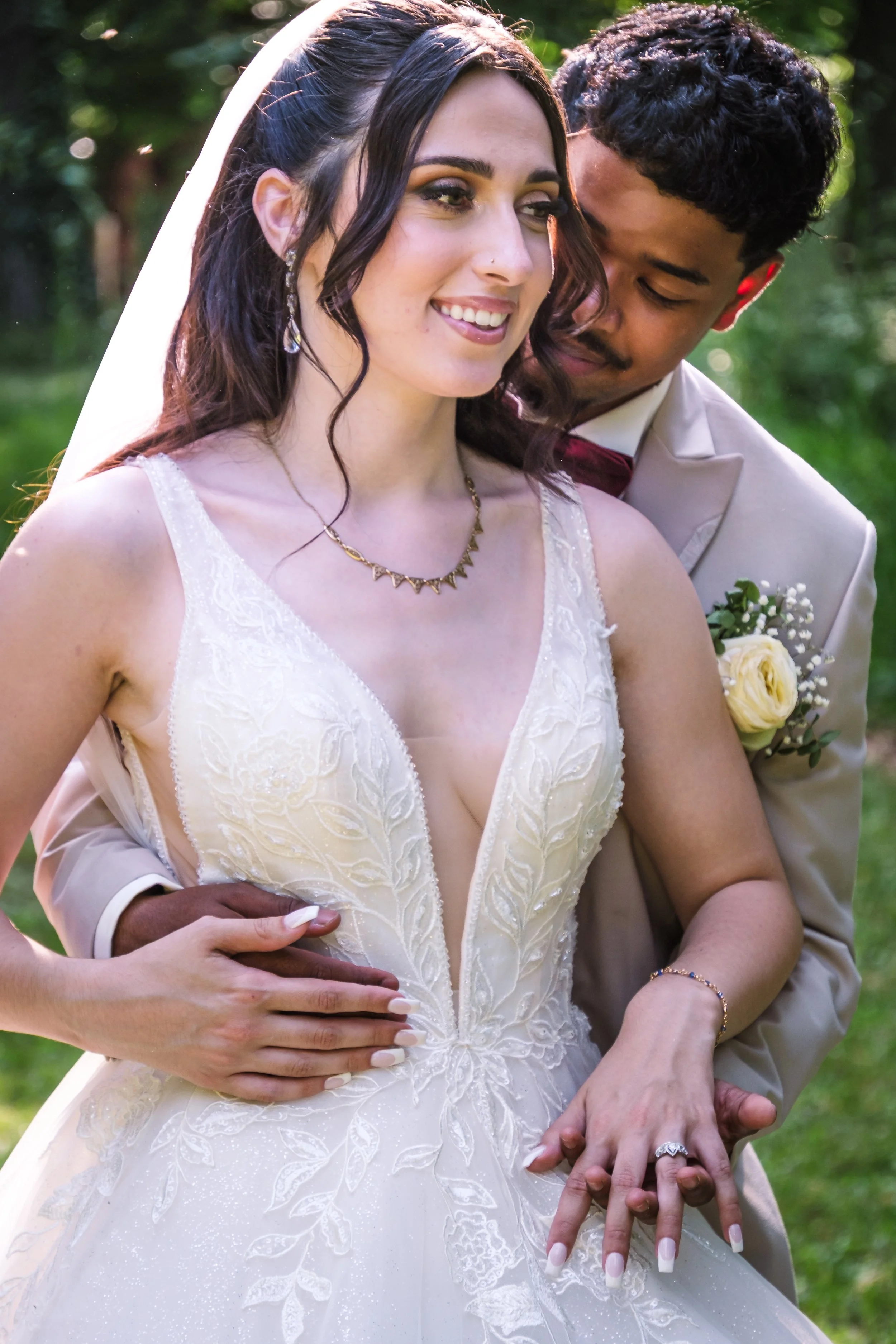 Un couple de mariés lors d'un mariage en plein air. La mariée porte une robe blanche avec des broderies florales, un collier doré et un voile blanc. Le marié porte un costume beige avec une chemise blanche, une cravate rouge et une boutonnière. Ils s