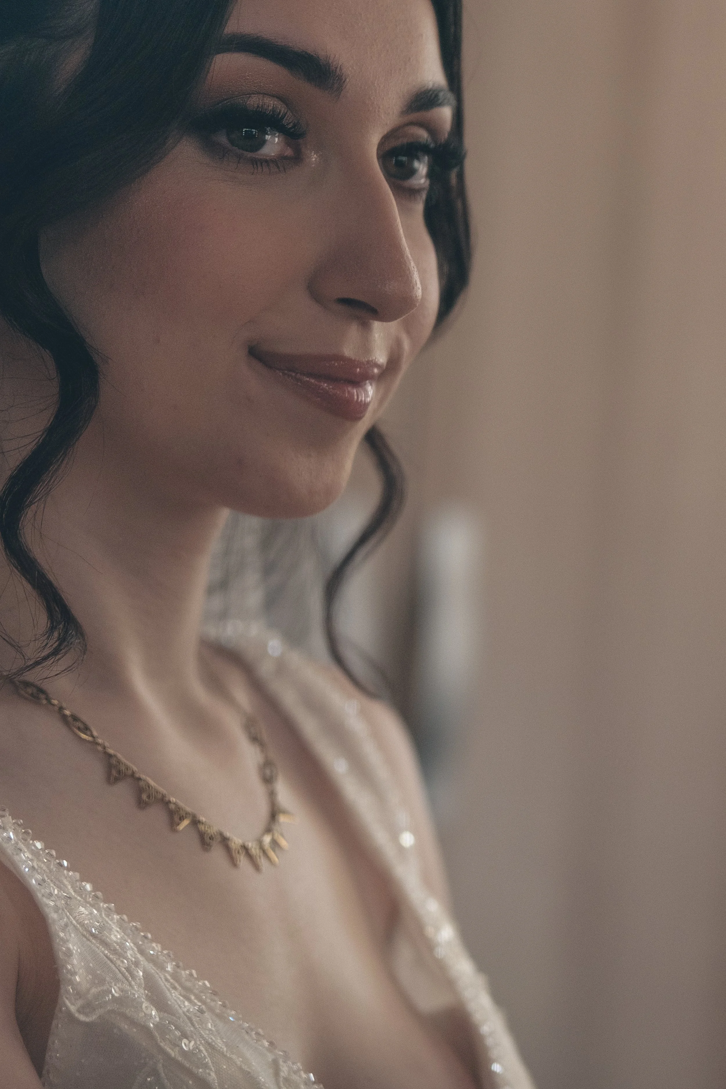 Une femme souriante avec des cheveux bouclés noirs portant un collier en or et une robe de mariage avec des détails en dentelle et perles.