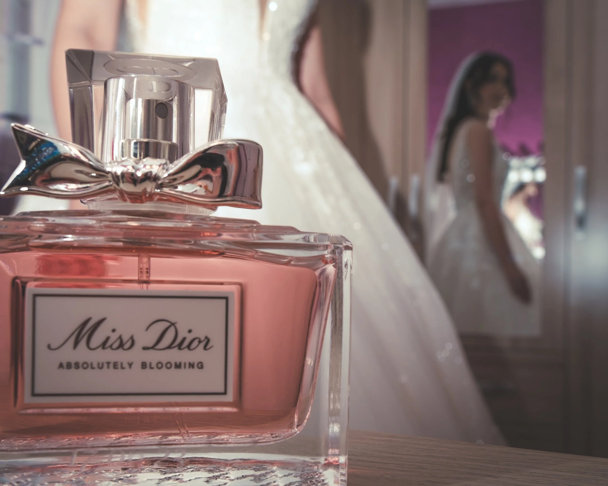 Un flacon de parfum Miss Dior Absolutely Blooming en premier plan, avec une robe de mariée en arrière-plan et un miroir dans une pièce lumineuse.