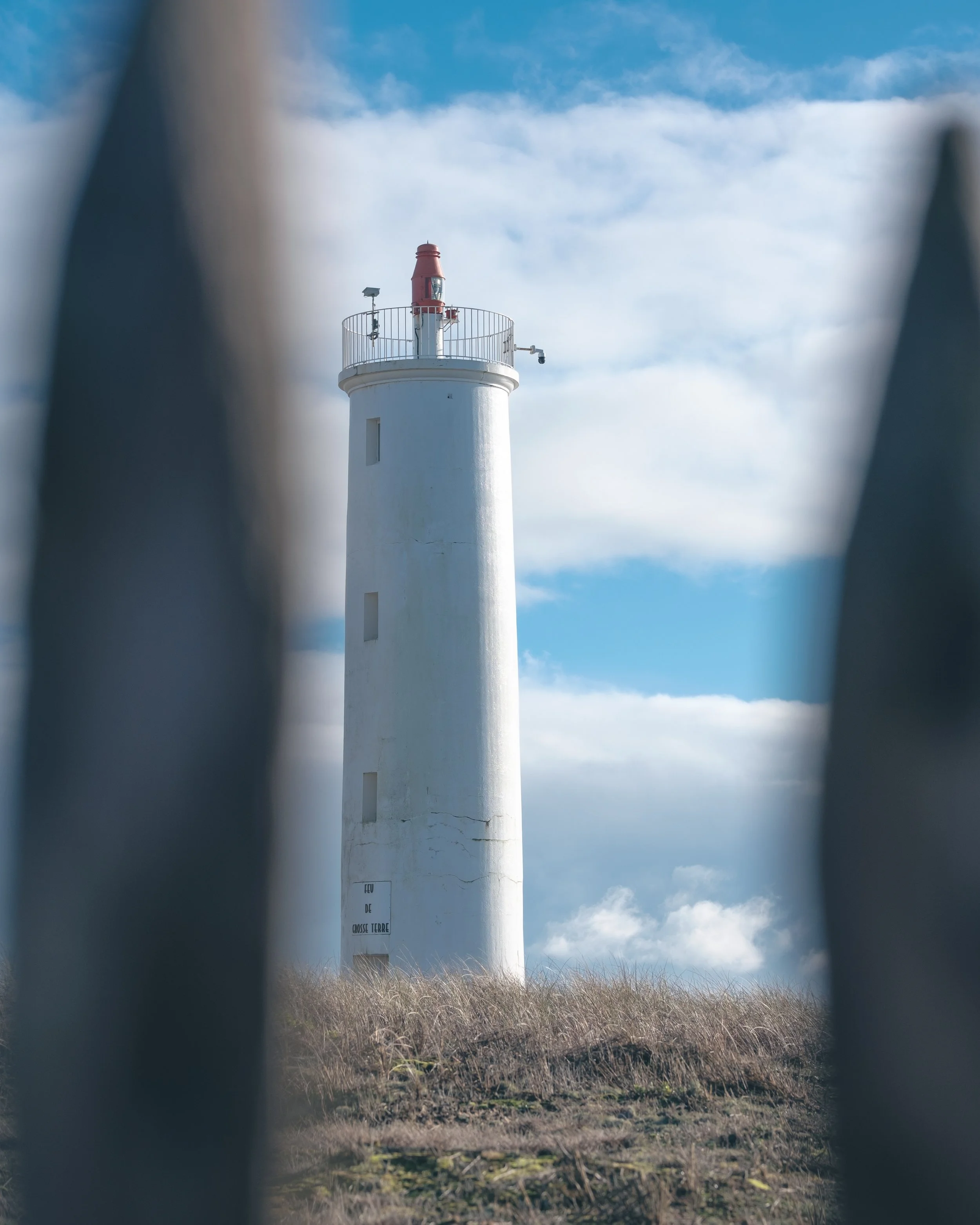 Phare blanc avec railing au sommet, vu à travers une clôture métallique en gros plan, ciel bleu et nuages en arrière-plan.