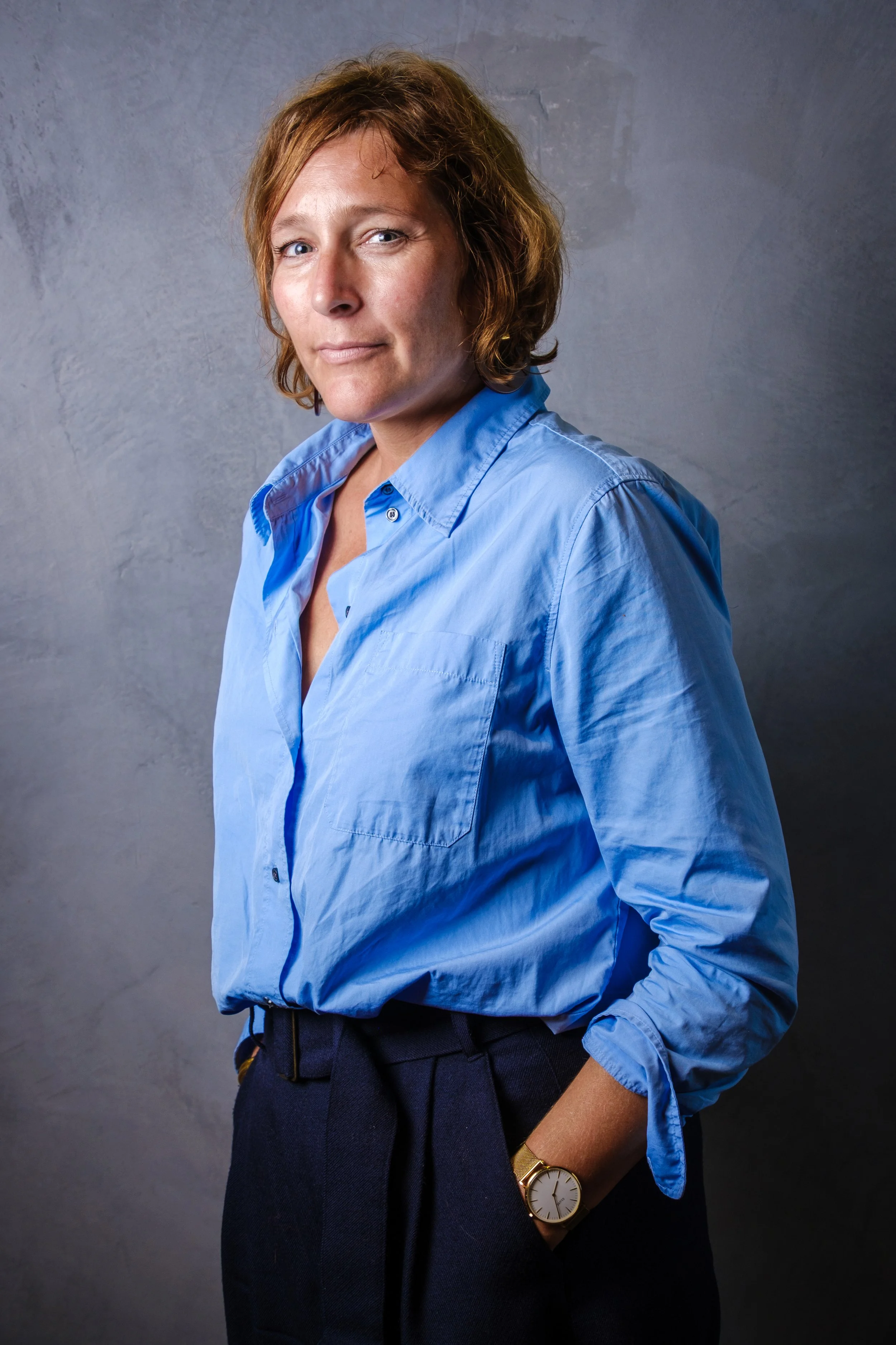 Une femme aux cheveux bruns ondulés portant une chemise bleue et un pantalon noir, avec sa main dans la poche, regardant vers l'objectif, sur un fond gris.