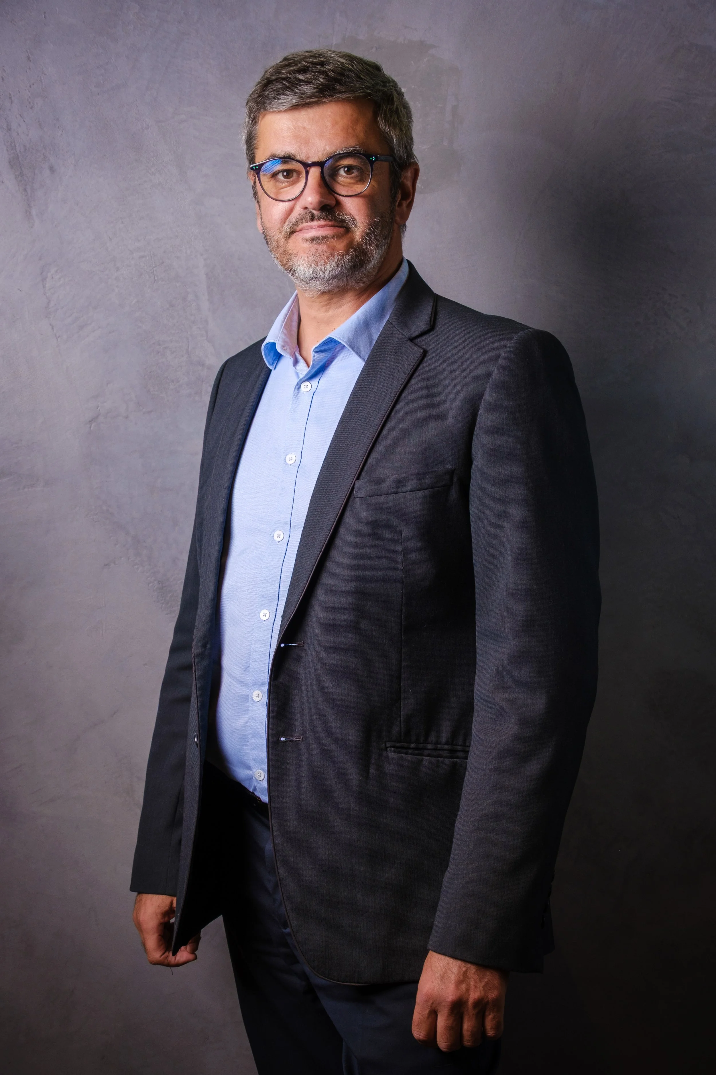 Homme d'âge moyen portant un costume noir et une chemise bleue, portant des lunettes, posant devant un mur gris.