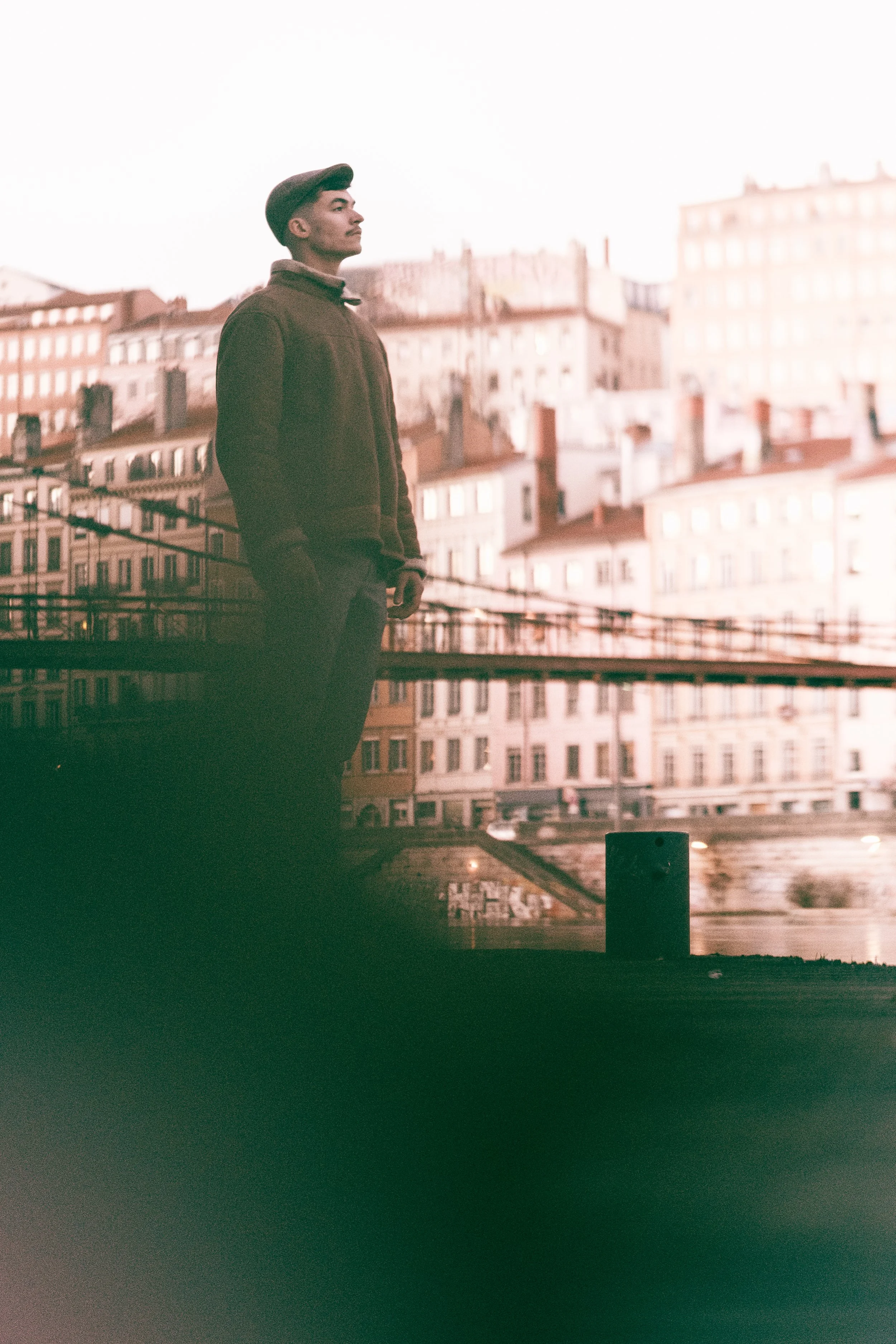 Un homme debout sur un pont à côté d'une rivière, avec des bâtiments résidentiels en arrière-plan, lors d'une journée nuageuse.
