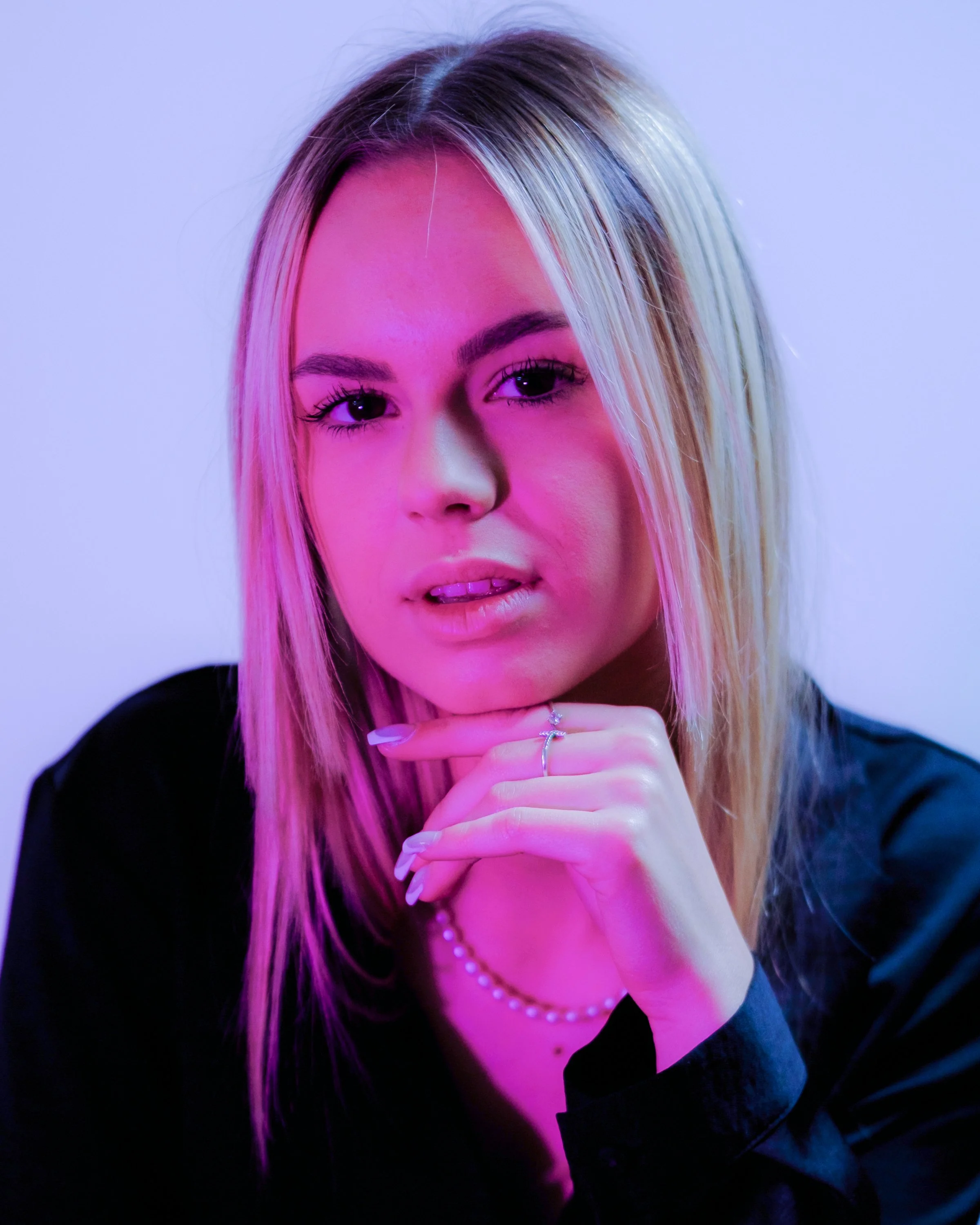 Jeune femme avec cheveux blonds, portant une veste noire, posant avec la main sous le menton, en lumière rose et violette.