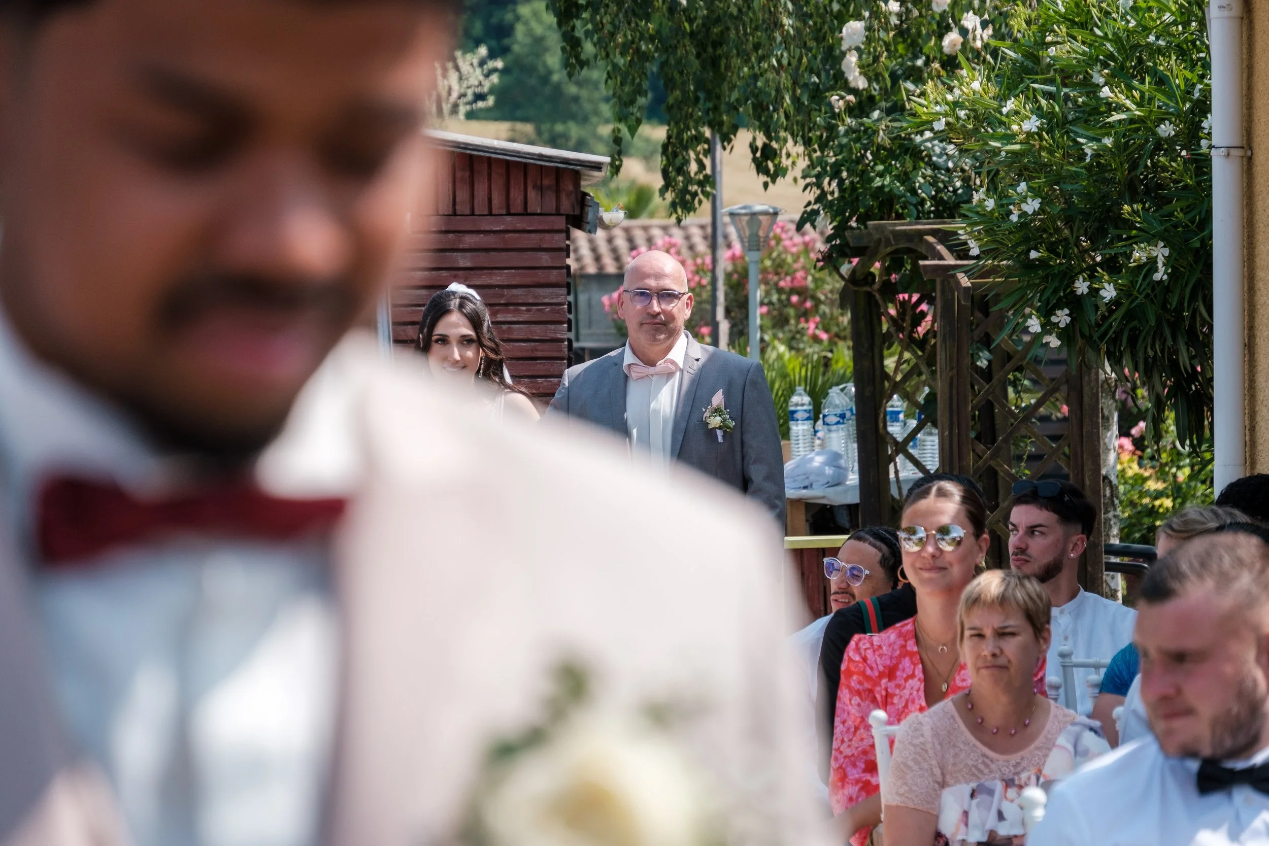 Le marié en premier plan flou, en costume blanc avec un nœud papillon rouge, lors d'un mariage en extérieur, avec des invités assis à l'arrière et une homme en costume gris dans le fond, souriant, entouré de fleurs et de végétation.