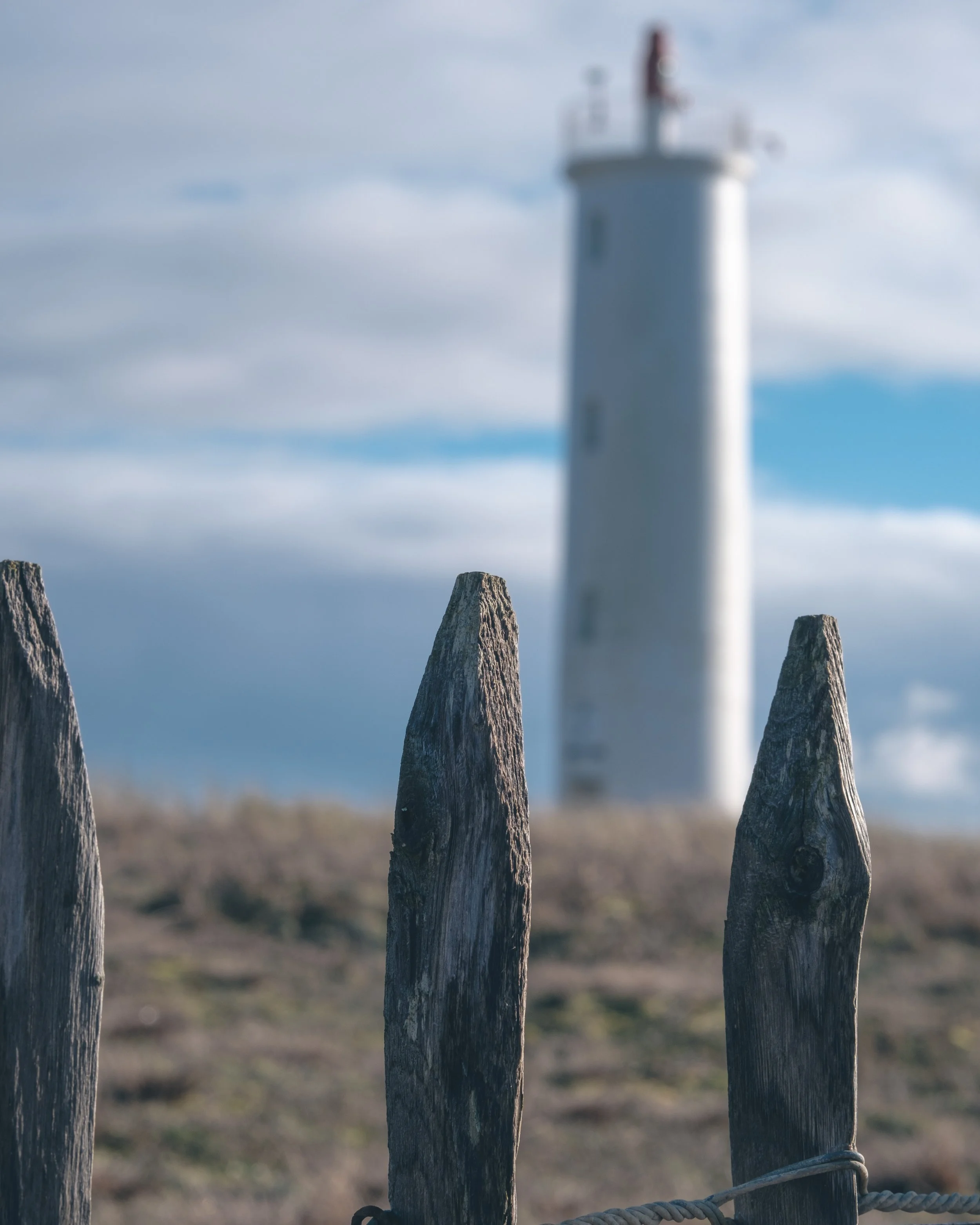 Poteaux en bois sur une clôture en corde, avec un phare en arrière-plan sous un ciel nuageux.