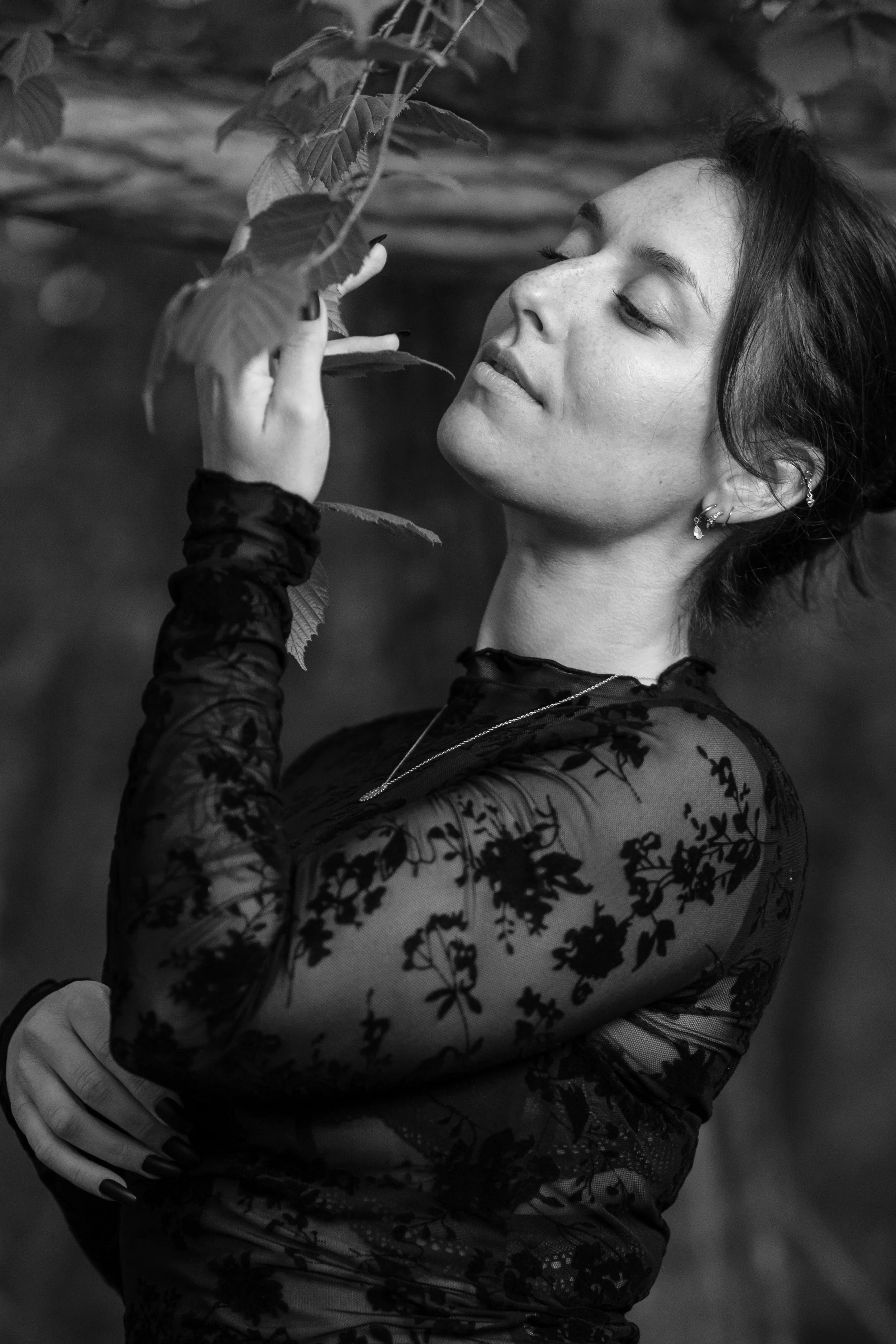 Une femme en vêtement noir en dentelle avec des fleurs, touchant des feuilles d'arbre.