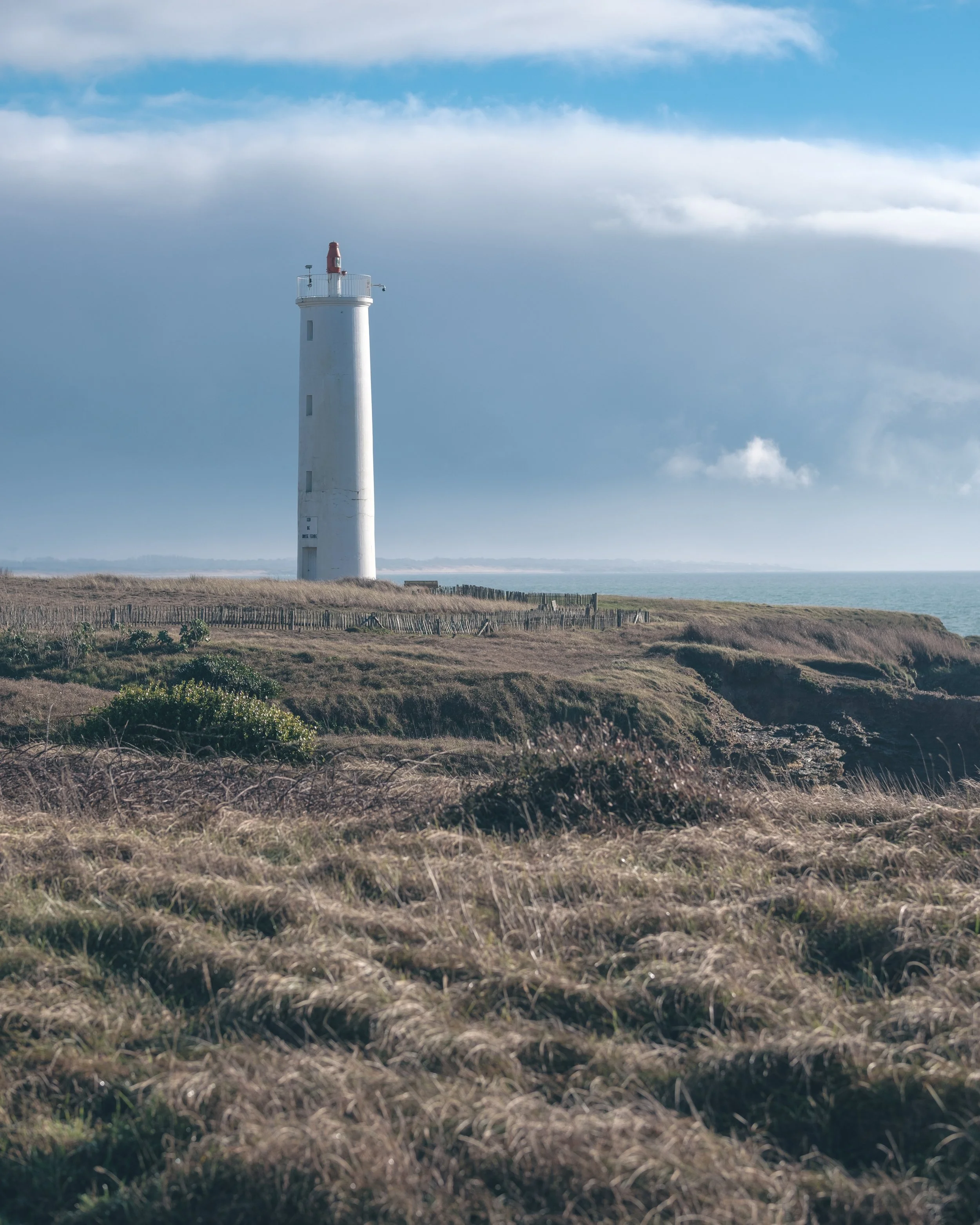 Phare blanc sur une côte rocheuse avec des herbes sauvages, ciel nuageux, en arrière-plan la mer.