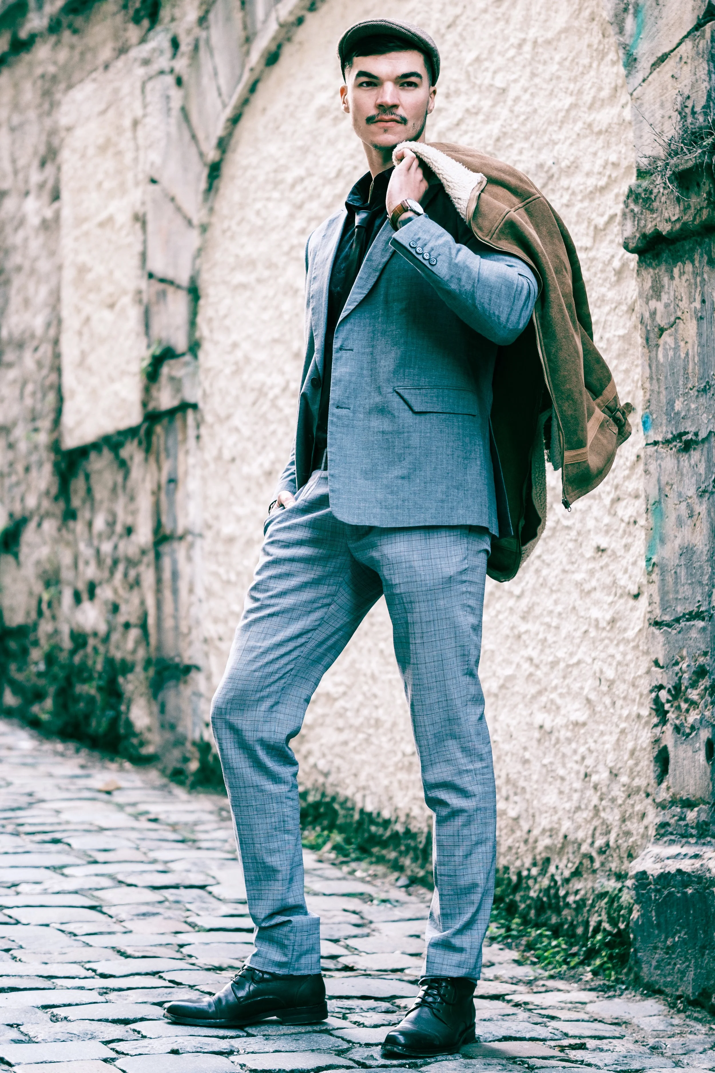 Homme en costume gris portant un sac à dos, debout sur un trottoir pavé, près d'un mur en pierre.