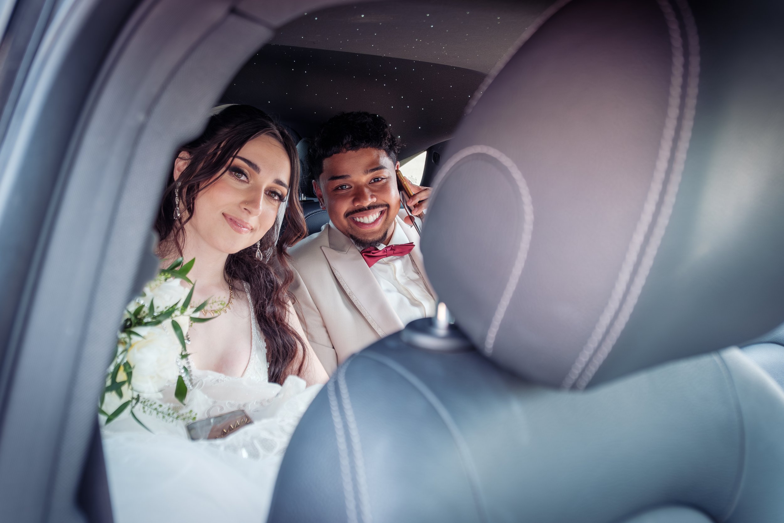 Un couple de jeunes mariés assis à l'arrière d'une voiture, souriant à la caméra, avec la mariée en robe blanche et le marié en costume clair avec nœud papillon, lors de leur mariage.
