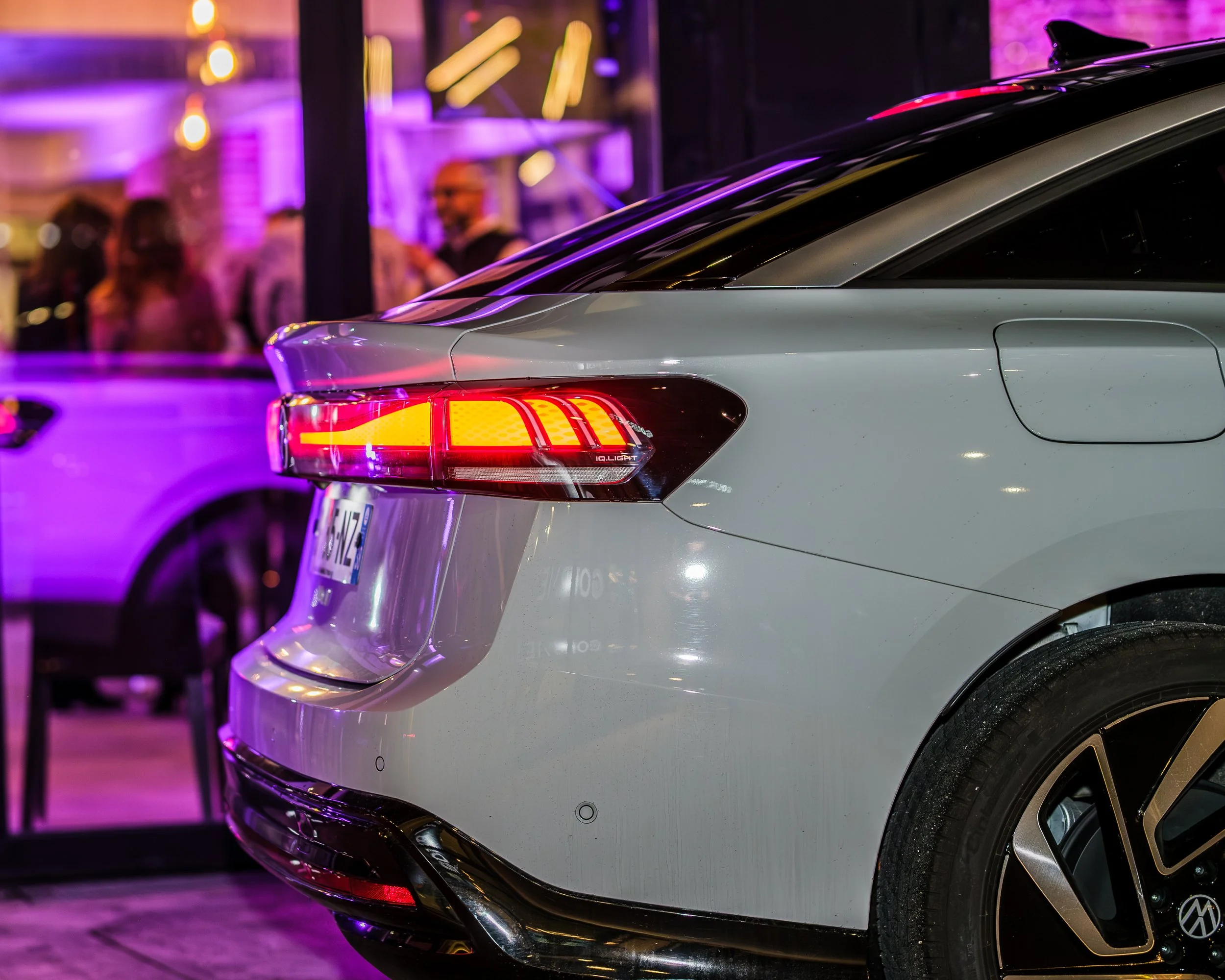 Une voiture blanche avec des lumières rouges allumées, vue de profil arrière, dans un environnement urbain nocturne avec des personnes floues en arrière-plan.