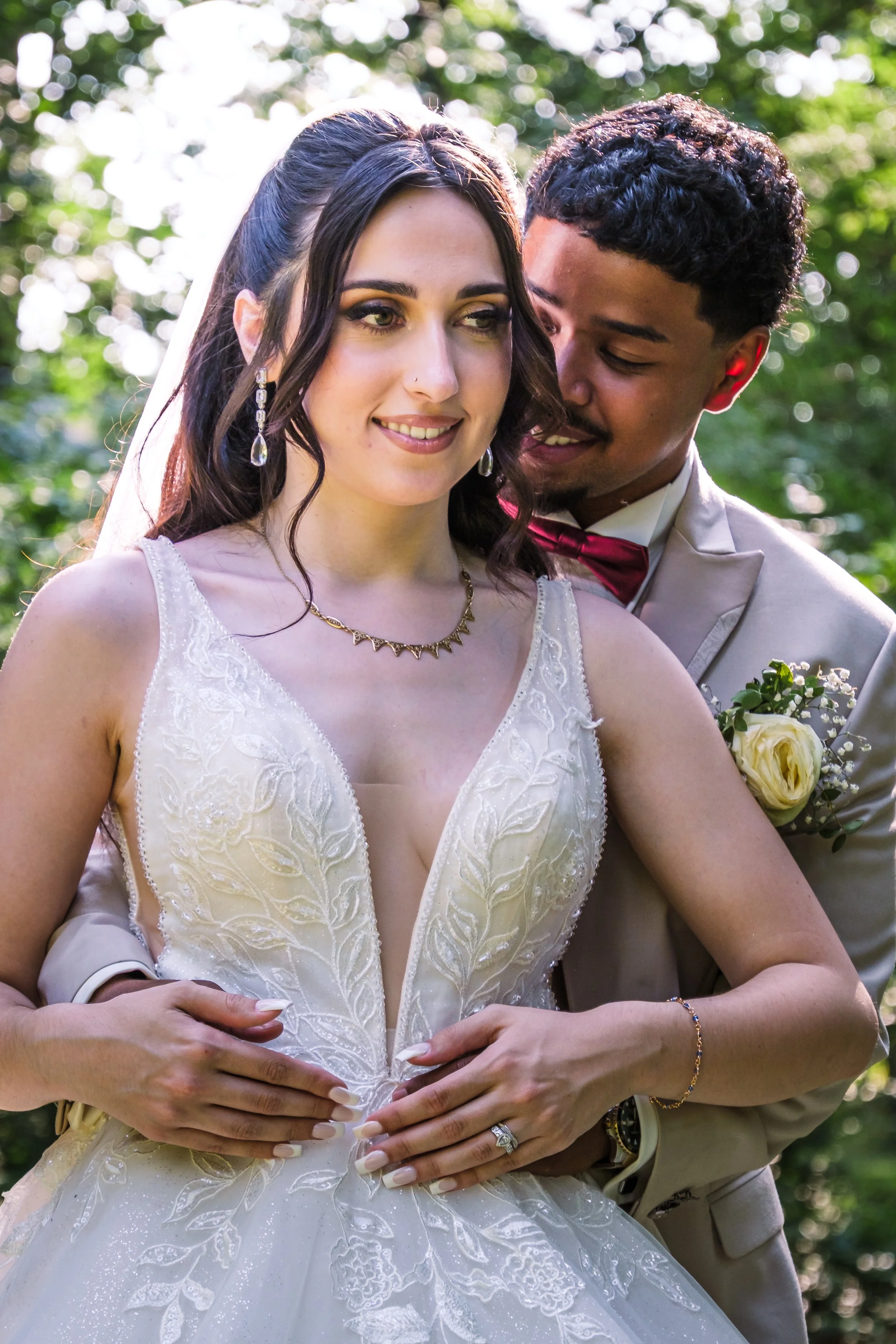 Un couple de mariés s'embrasse dans un cadre verdoyant, la mariée porte une robe blanche délicate avec des détails floraux, des boucles d'oreilles pendantes, et un collier, tandis que le marié porte un costume beige avec une boutonnière blanche.