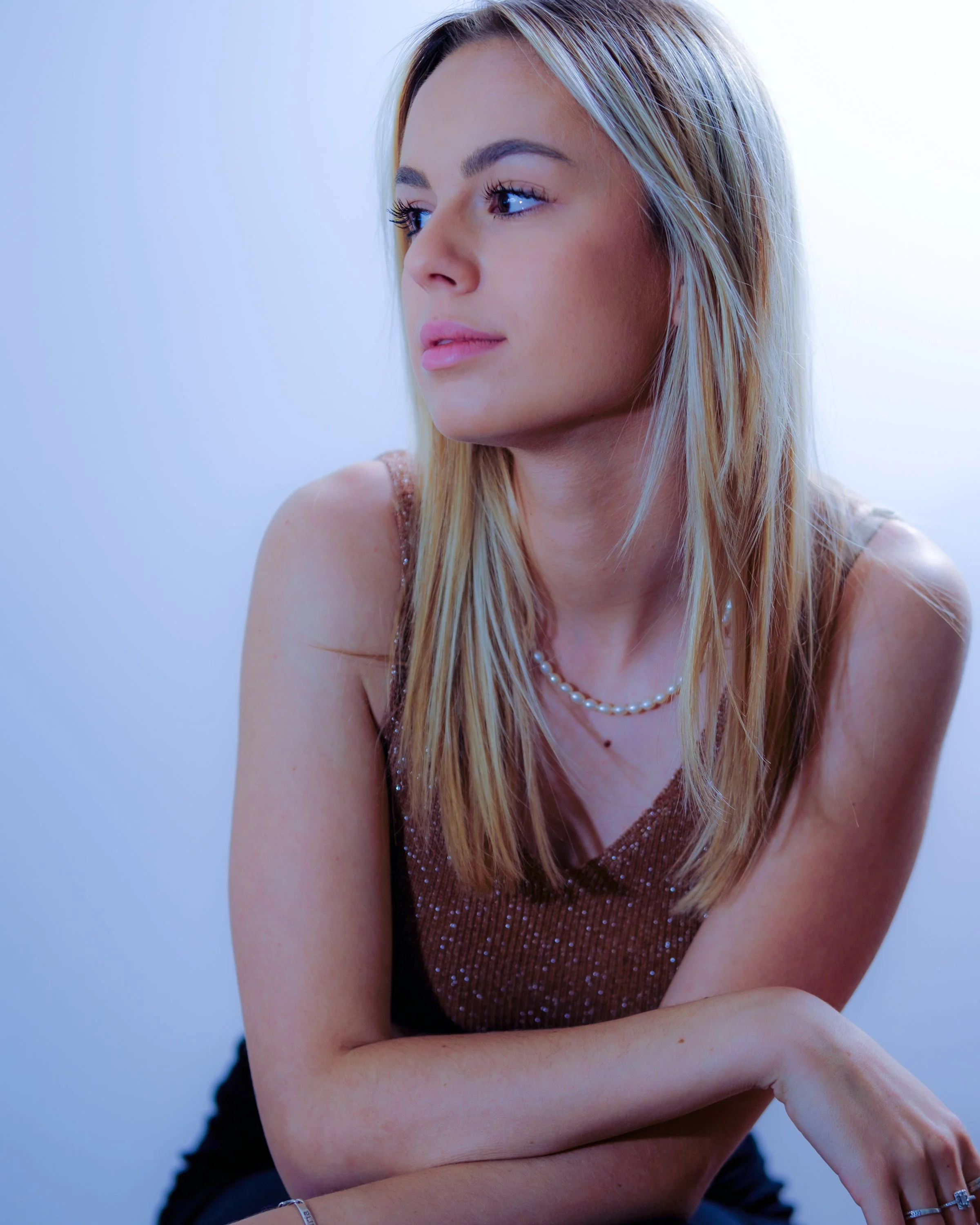 Jeune femme aux cheveux blonds, portant un haut pailleté marron, un collier de perles, et un bracelet, assise en regardant à gauche.
