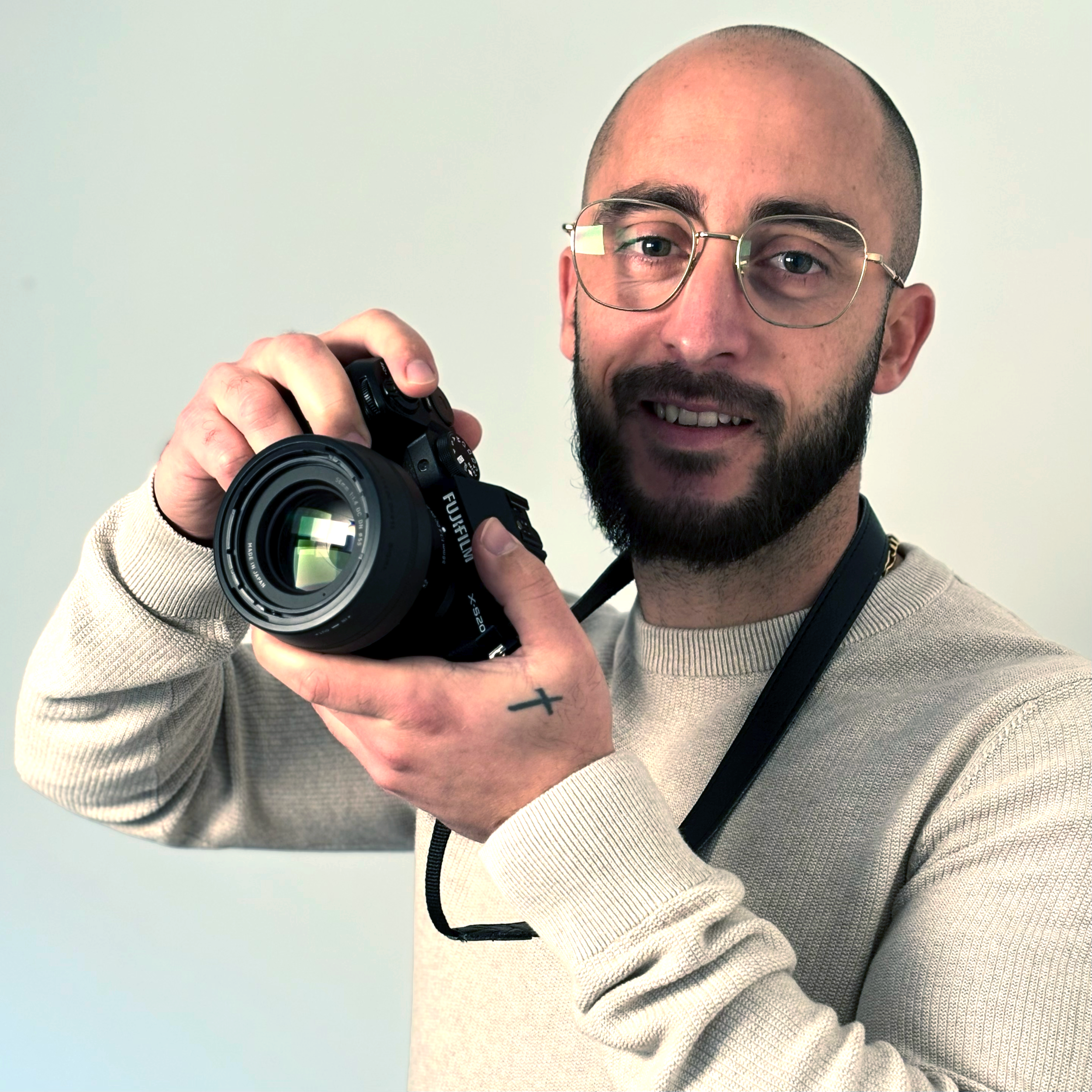 Un homme portant des lunettes, avec une barbe, tenant un appareil photo face à lui, souriant, portant un sweat beige et une caméra autour du cou.