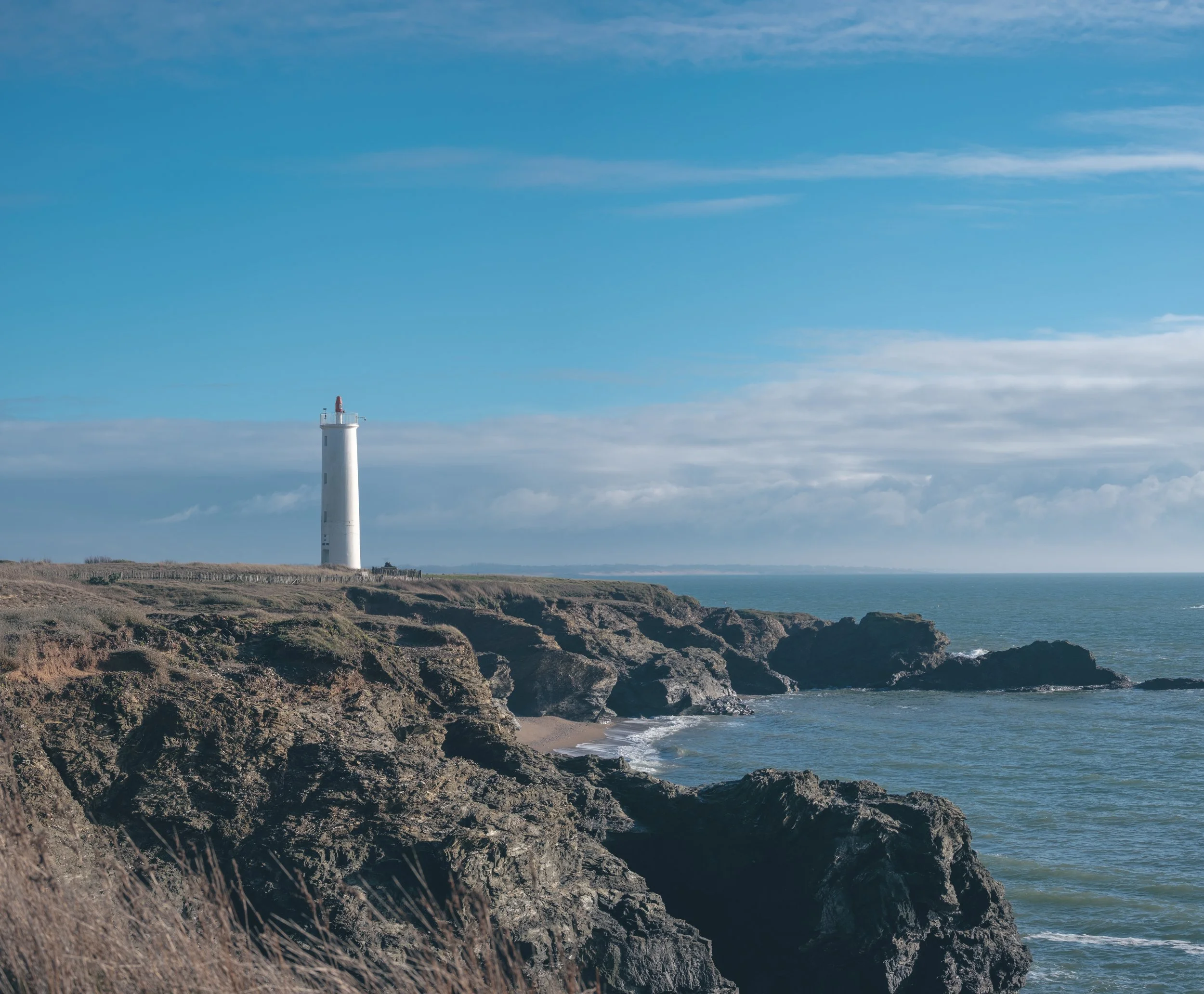 Phare blanc sur des falaises rocheuses au bord de la mer sous un ciel partiellement nuageux.