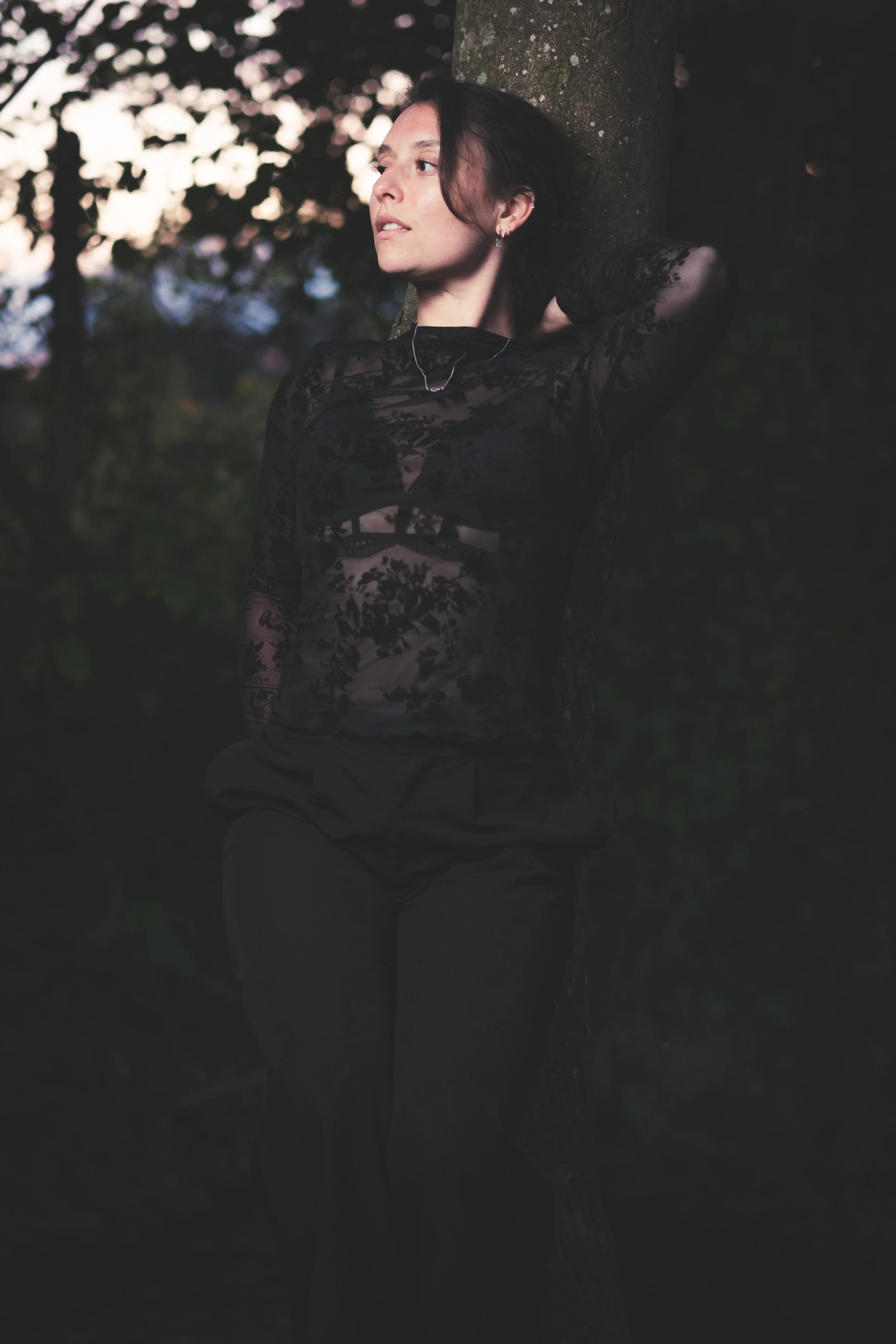 Une femme en vêtements noirs pose à côté d'un arbre dans un environnement naturel, avec un arrière-plan flou de feuillages et de lumière naturelle