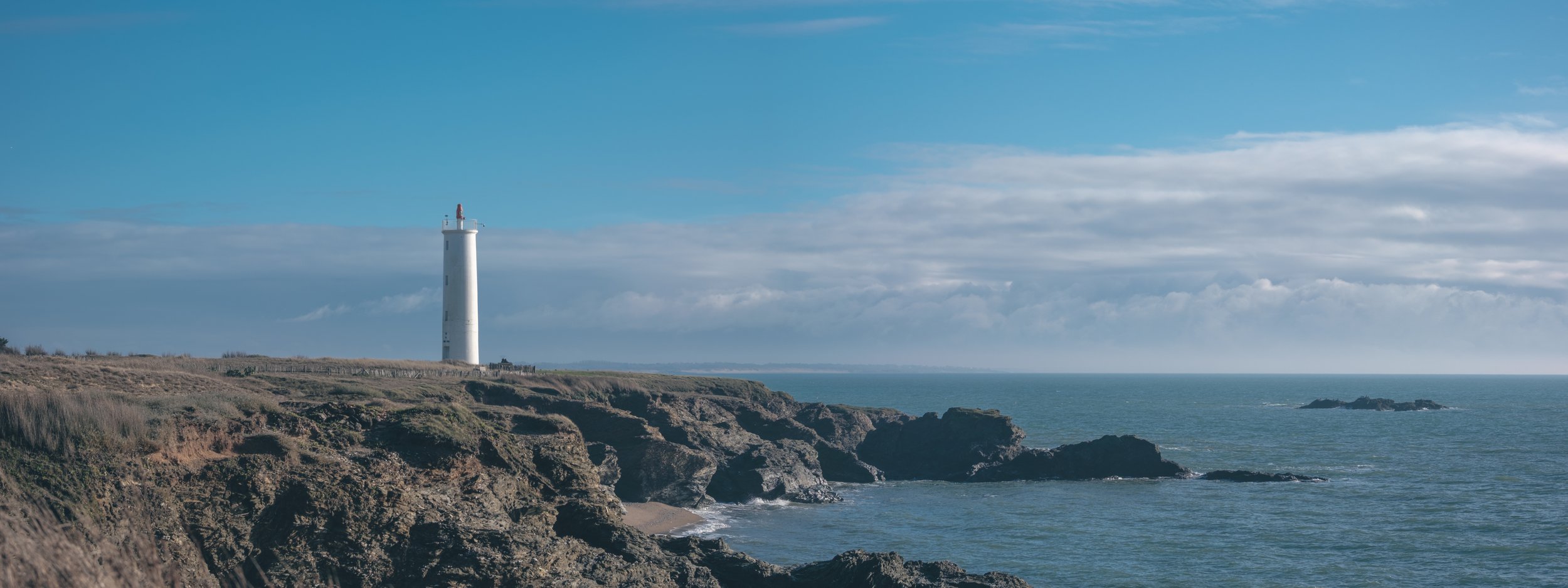 Phare blanc sur une falaise rocheuse au bord de la mer avec un ciel bleu et quelques nuages.