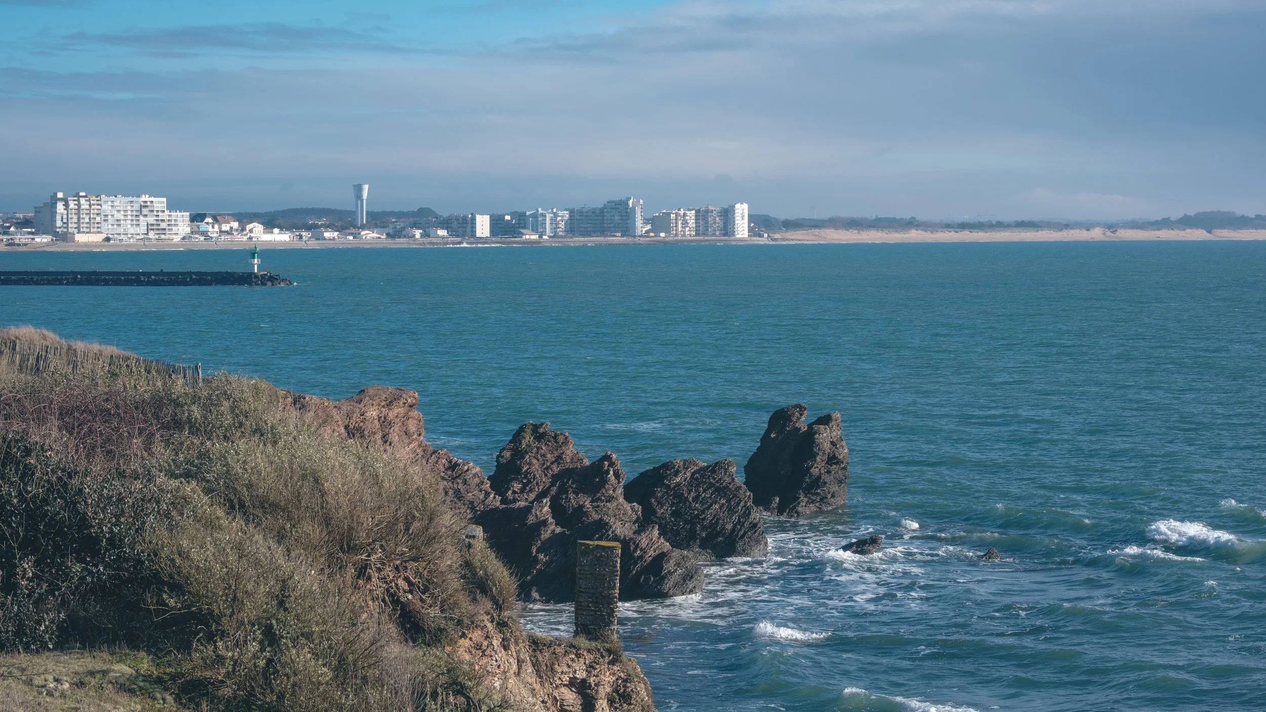 Vue d'une côte avec des rochers, de l'eau et des bâtiments en arrière-plan sous un ciel nuageux.