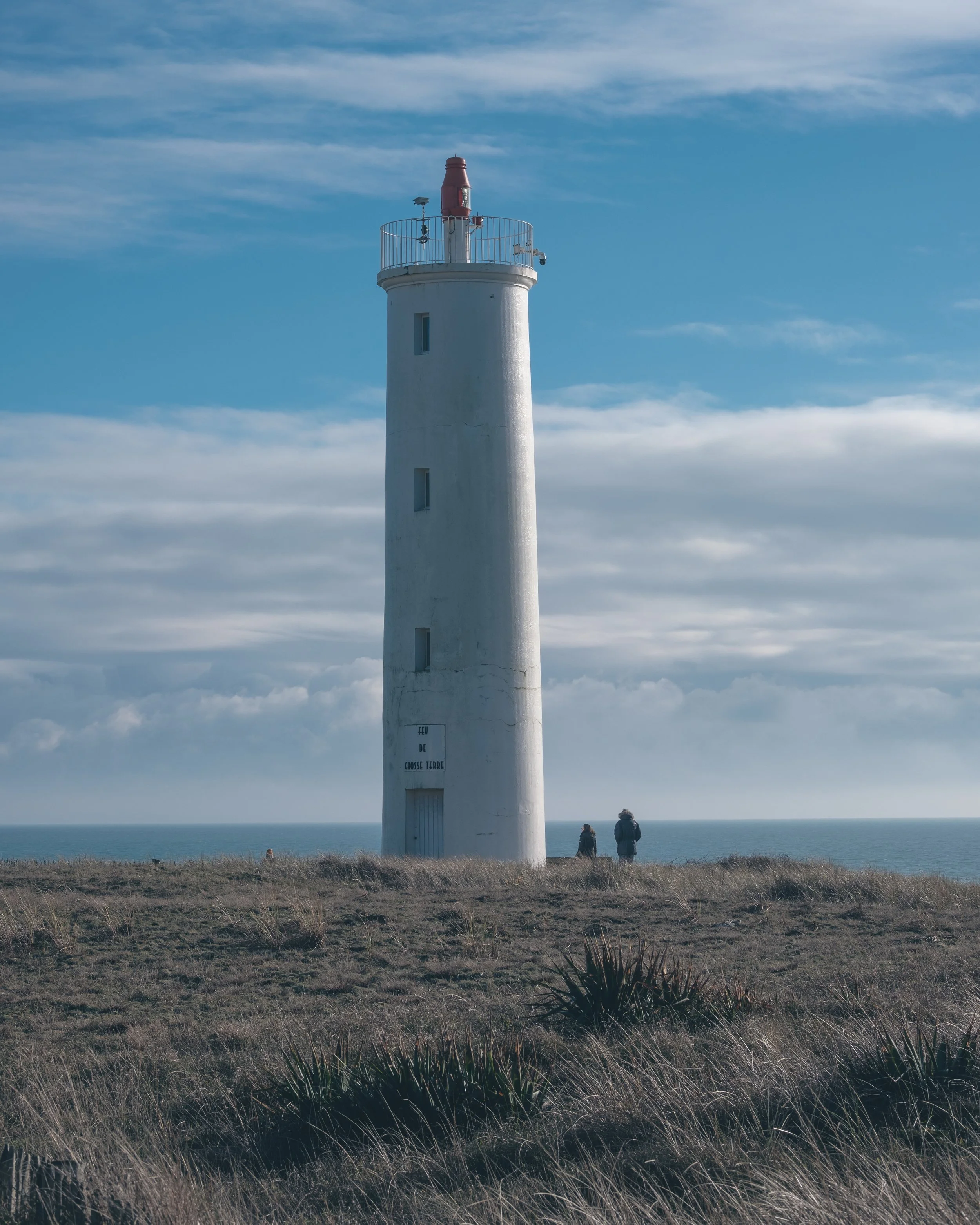 Phare blanc situé sur une côte avec deux personnes marchant à proximité, ciel nuageux et mer en arrière-plan.