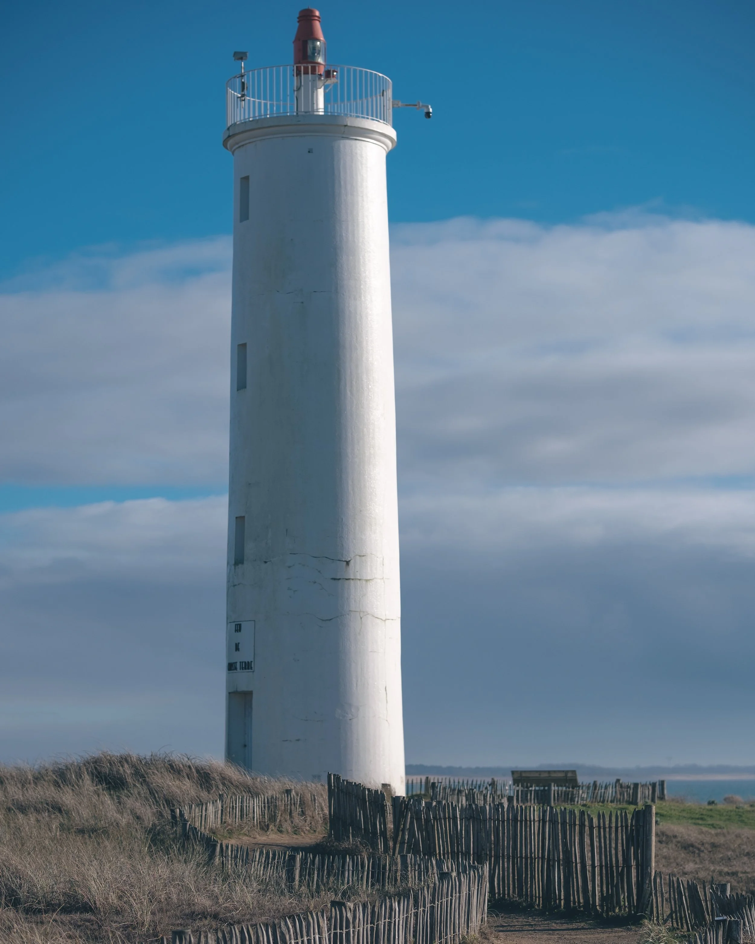 Phare blanc avec une rambarde rouge en haut, entouré de ciel bleu et de nuages, au bord de la mer, avec une clôture en bois sur le sentier.