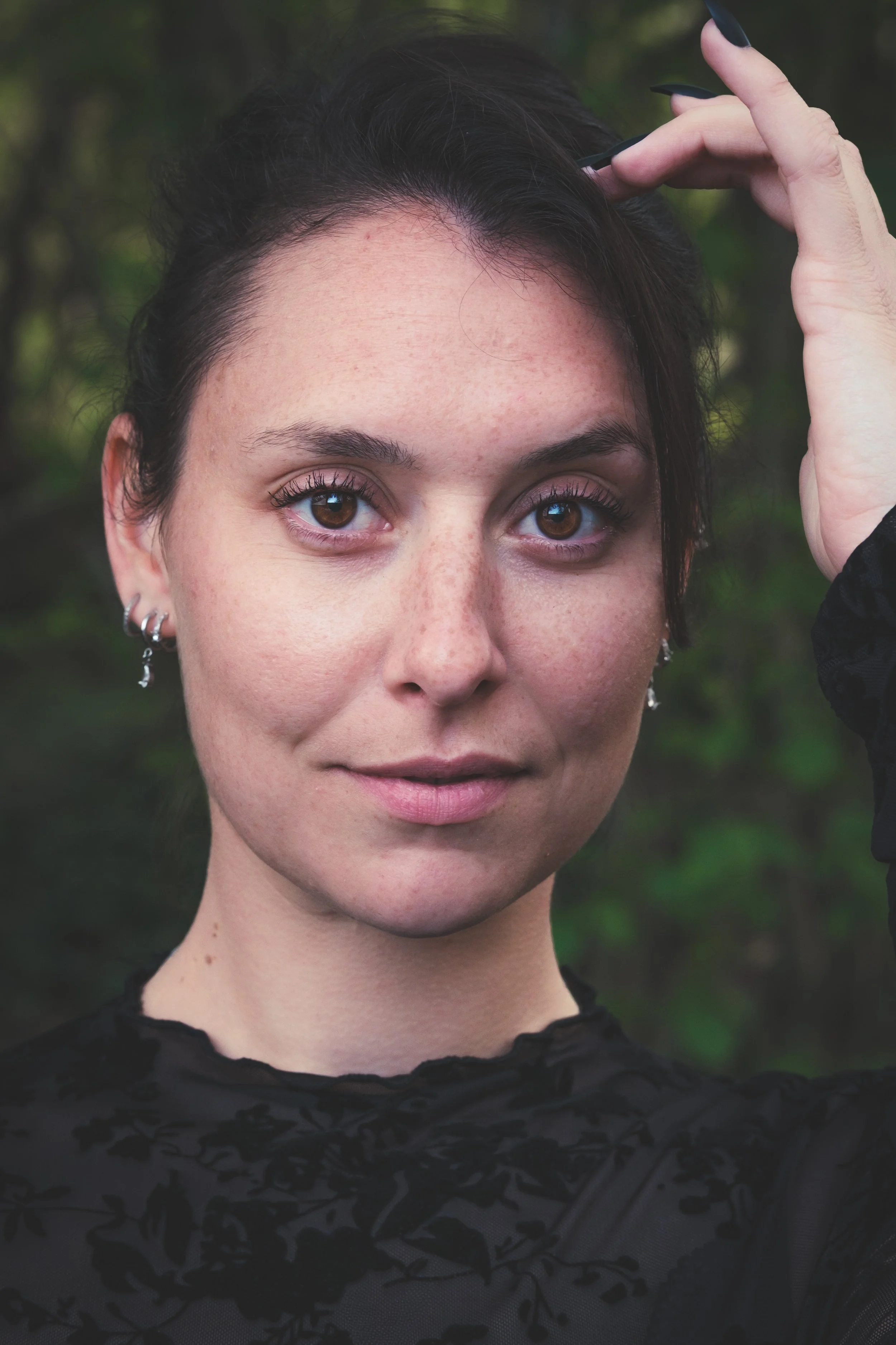 Jeune femme avec des yeux marron, des lèvres légèrement souriantes et des cheveux noirs, portant des boucles d'oreilles, en extérieur avec un fond flou de végétation.