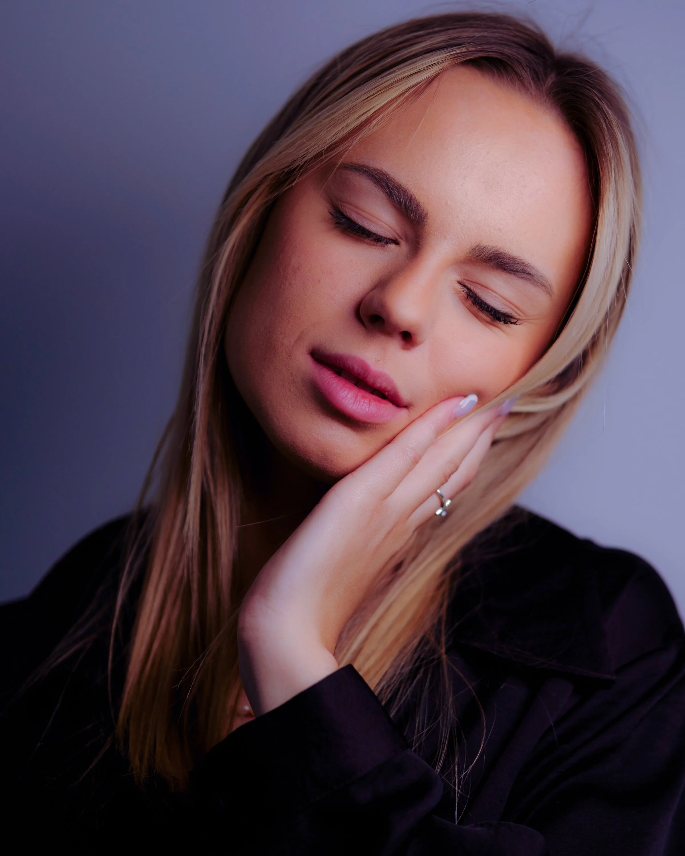 Jeune femme aux cheveux blonds et yeux fermés, touchant doucement sa joue avec ses doigts, portant un vêtement noir.