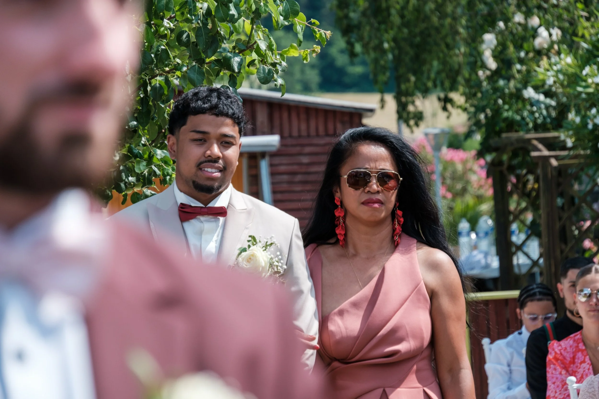 Deux personnes, un homme en costume blanc avec un nœud papillon rouge et une femme en robe rose avec des lunettes de soleil, lors d'un événement en extérieur.