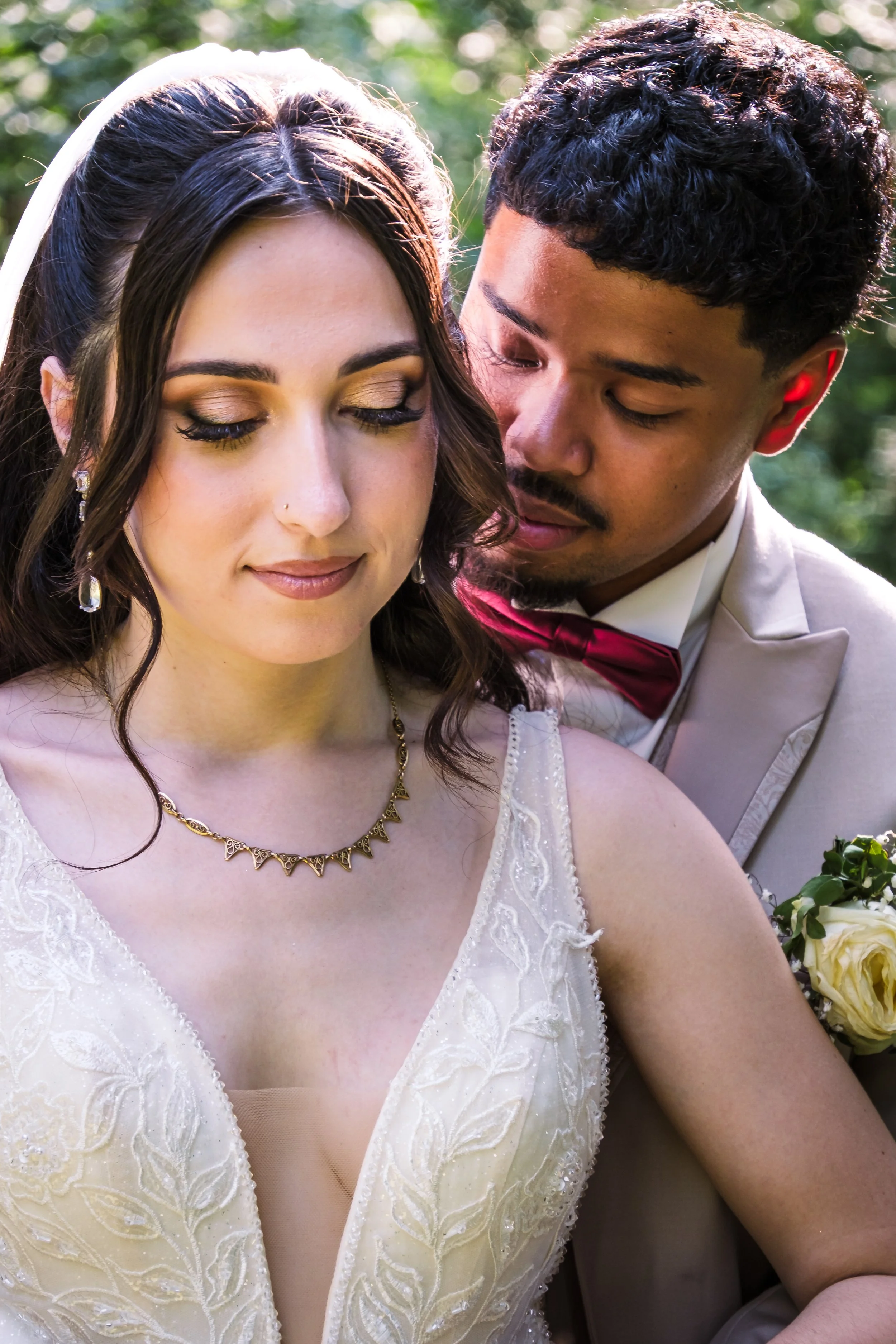 Un couple de mariés en extérieur, la femme porte une robe de mariage ivoire avec des détails en broderie, et le homme porte un costume gris clair avec une chemise blanche et un nœud papillon rouge. Ils sont proches, avec la femme qui a les yeux fermé