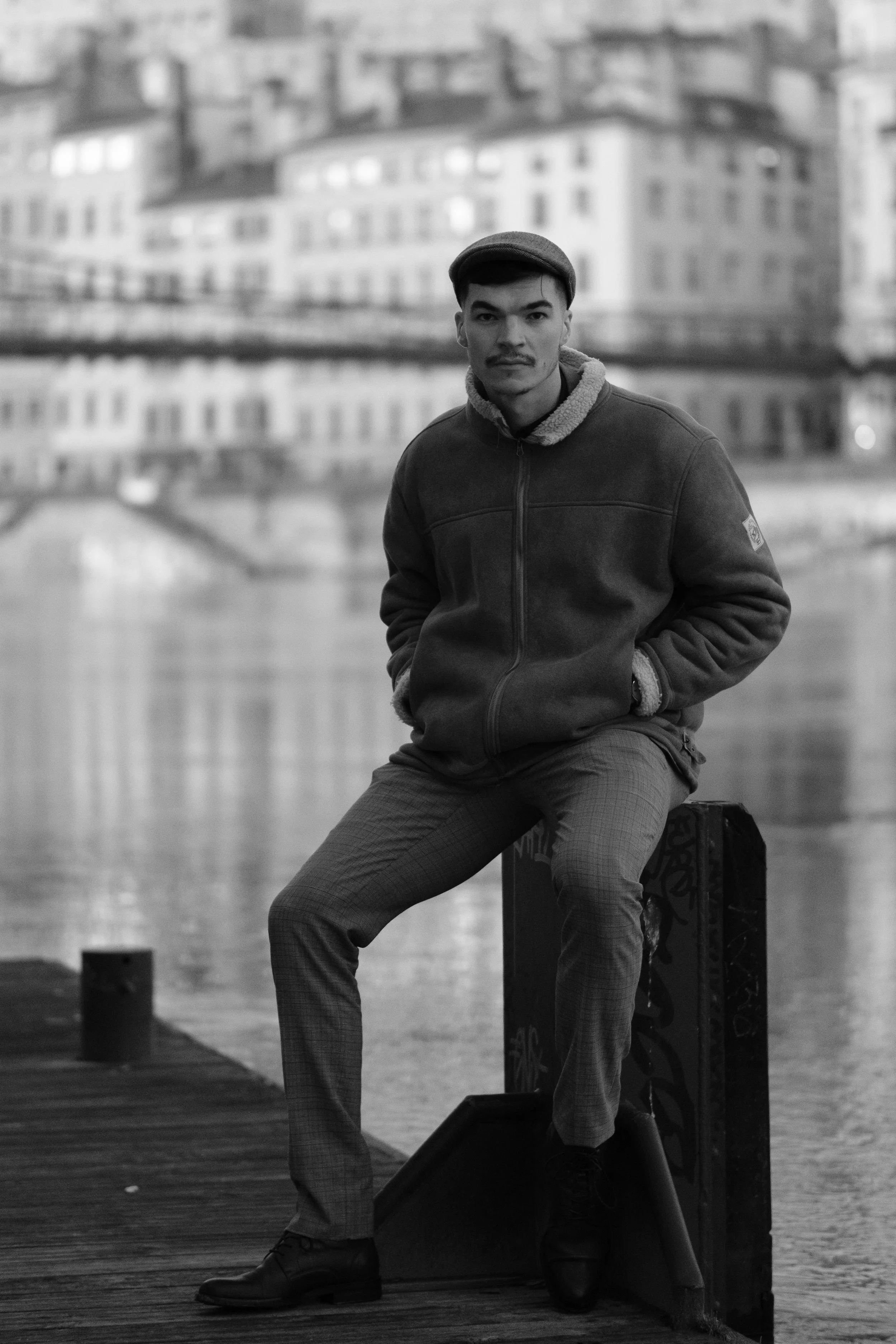 Un homme en veste et pantalon à carreaux, portant un bonnet, assis sur un dispositif près d'un plan d'eau dans un environnement urbain en noir et blanc.