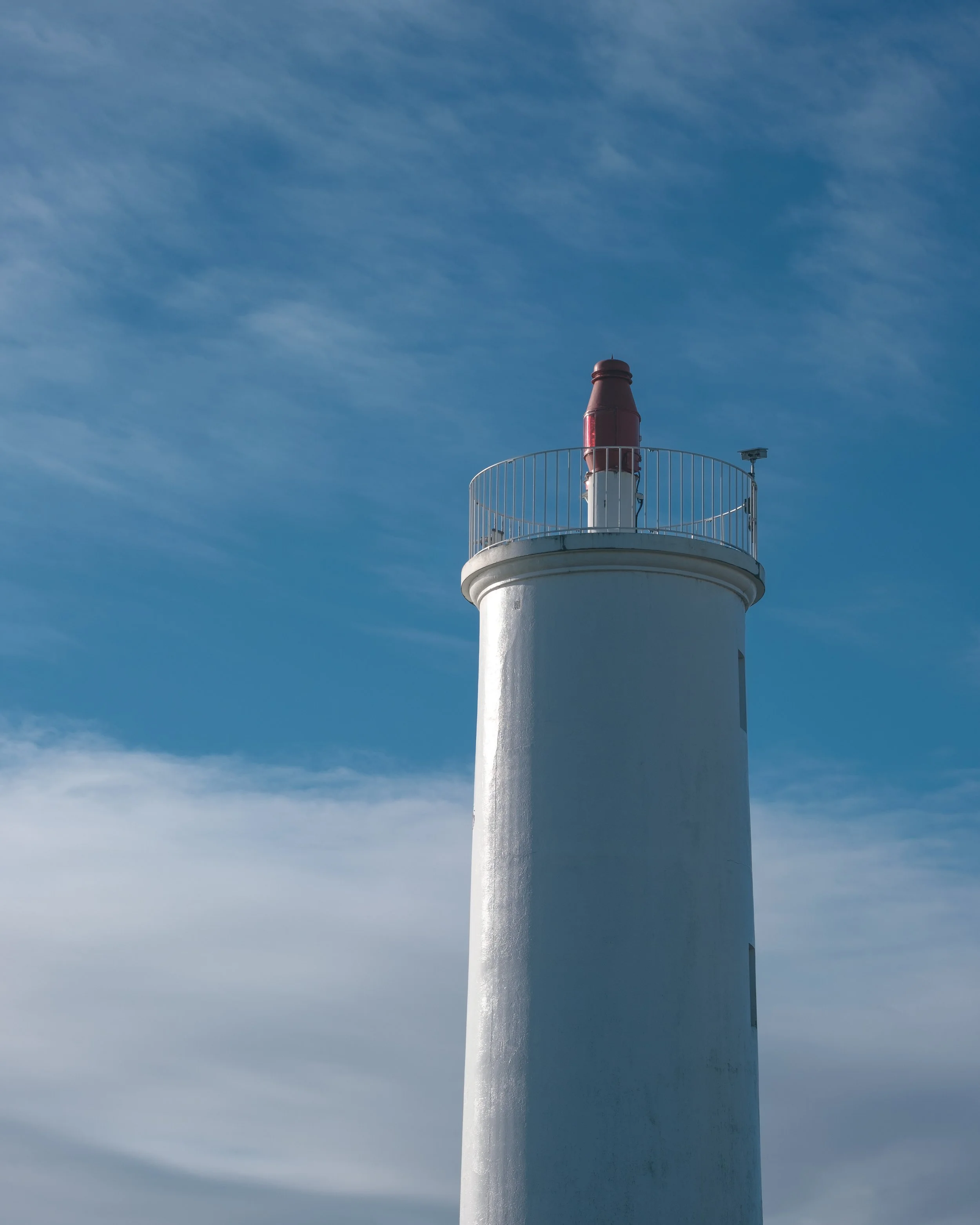 Phare blanc avec une plateforme en haut, contre un ciel bleu avec quelques nuages.