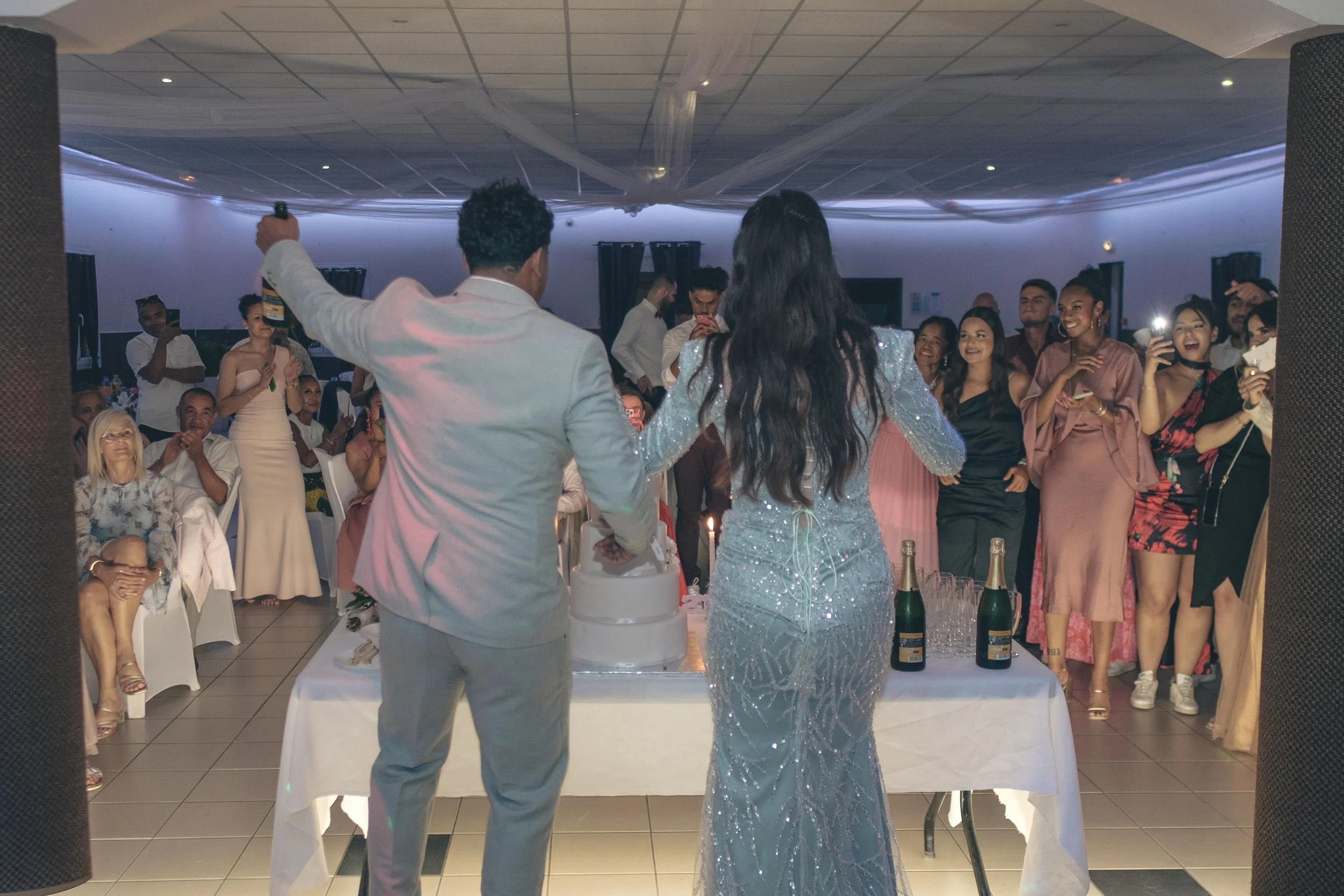 Une célébration de mariage avec un couple en robe de soirée et costume près d'un gâteau, entourés d'invités souriants dans une salle décorée, célébrant un moment festif.