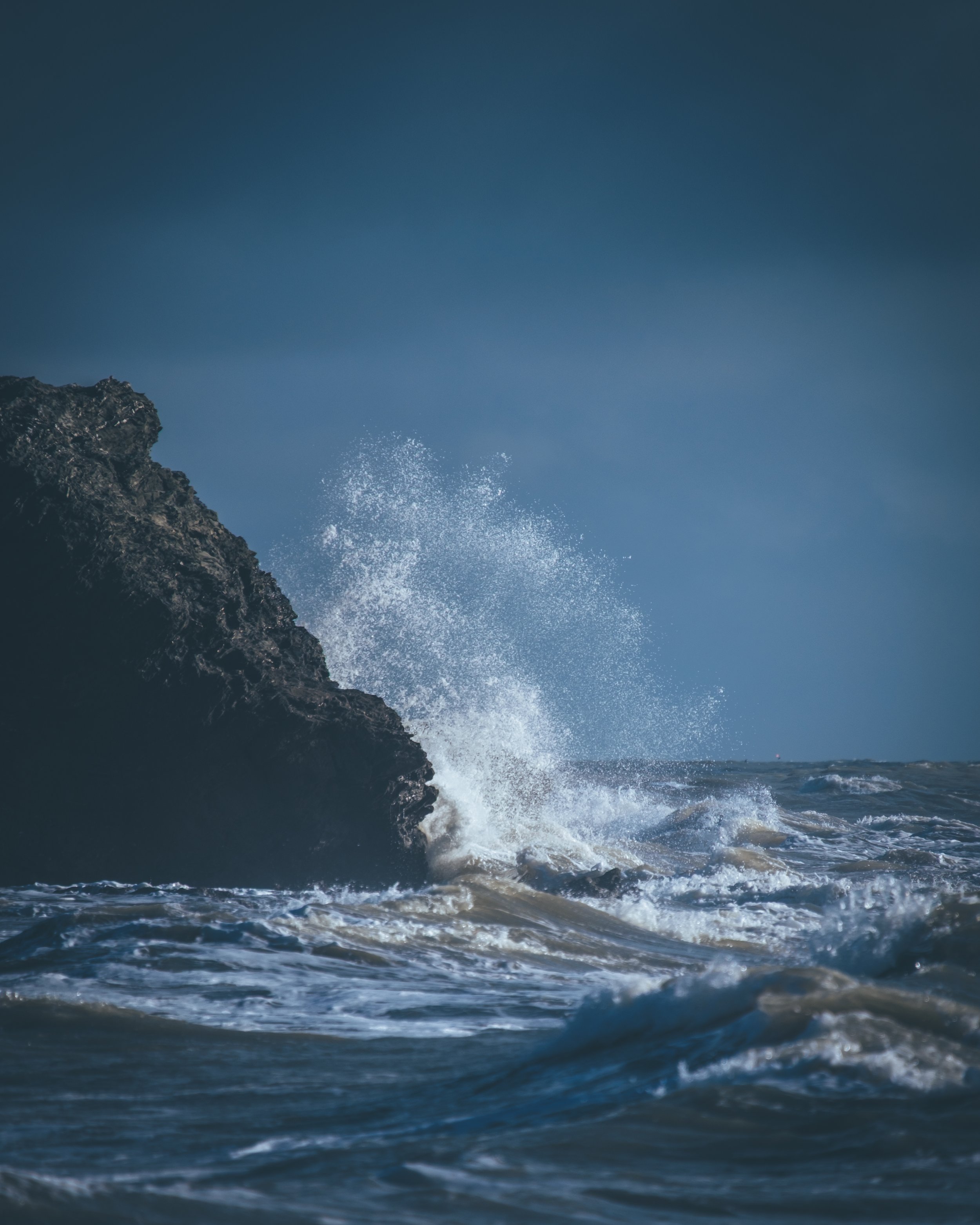 Vague de mer s'écrasant contre une roche escarpée en mer, avec un ciel nuageux en arrière-plan.
