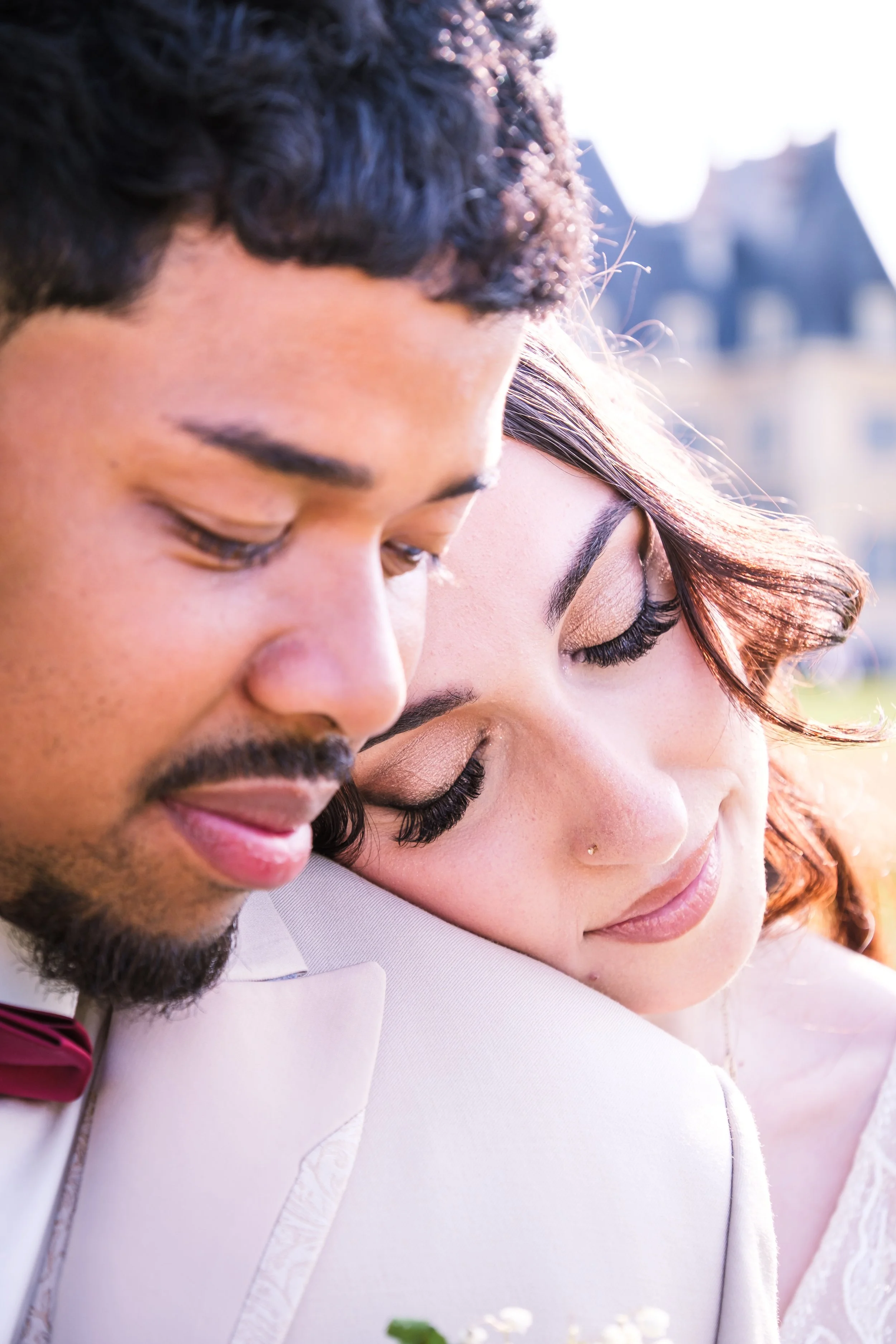 Un couple avec les yeux fermés, en tenue de mariage, partage un moment intime avec un fond flou d'architecture parisienne.